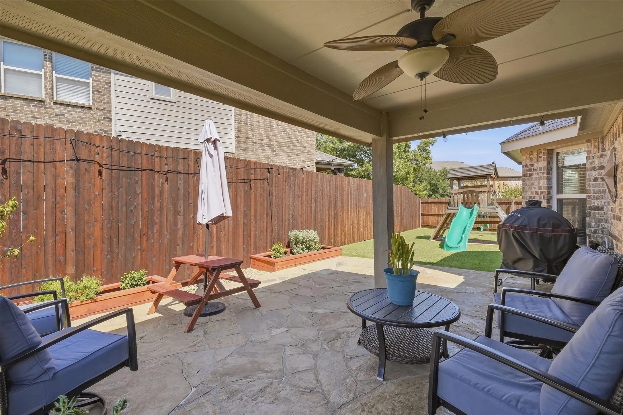 Covered flagstone patio complete with an outdoor ceiling fan, providing a shaded retreat overlooking the fenced lawn and playground area.