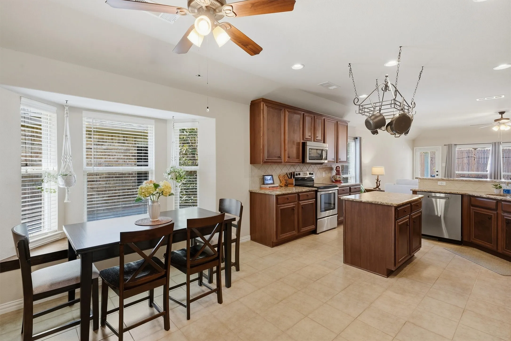 This sun-filled dining nook features a bay window, a lighted ceiling fan, and direct sightlines into the open-concept kitchen. The functional layout seamlessly connects everyday dining to the main kitchen island and living space beyond.