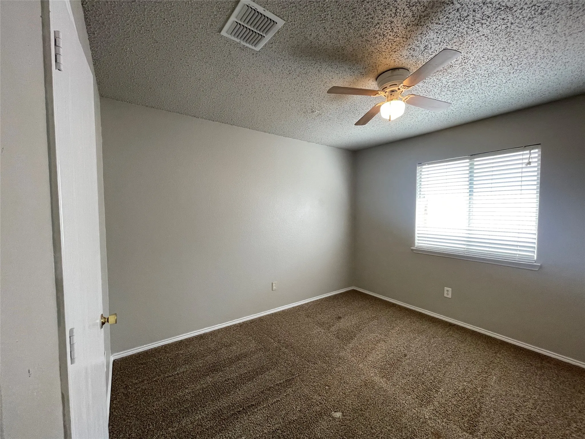 Neutral wall paint, brown carpeting, ceiling fan with integrated lighting, window with horizontal blinds, and a white door with brass hardware