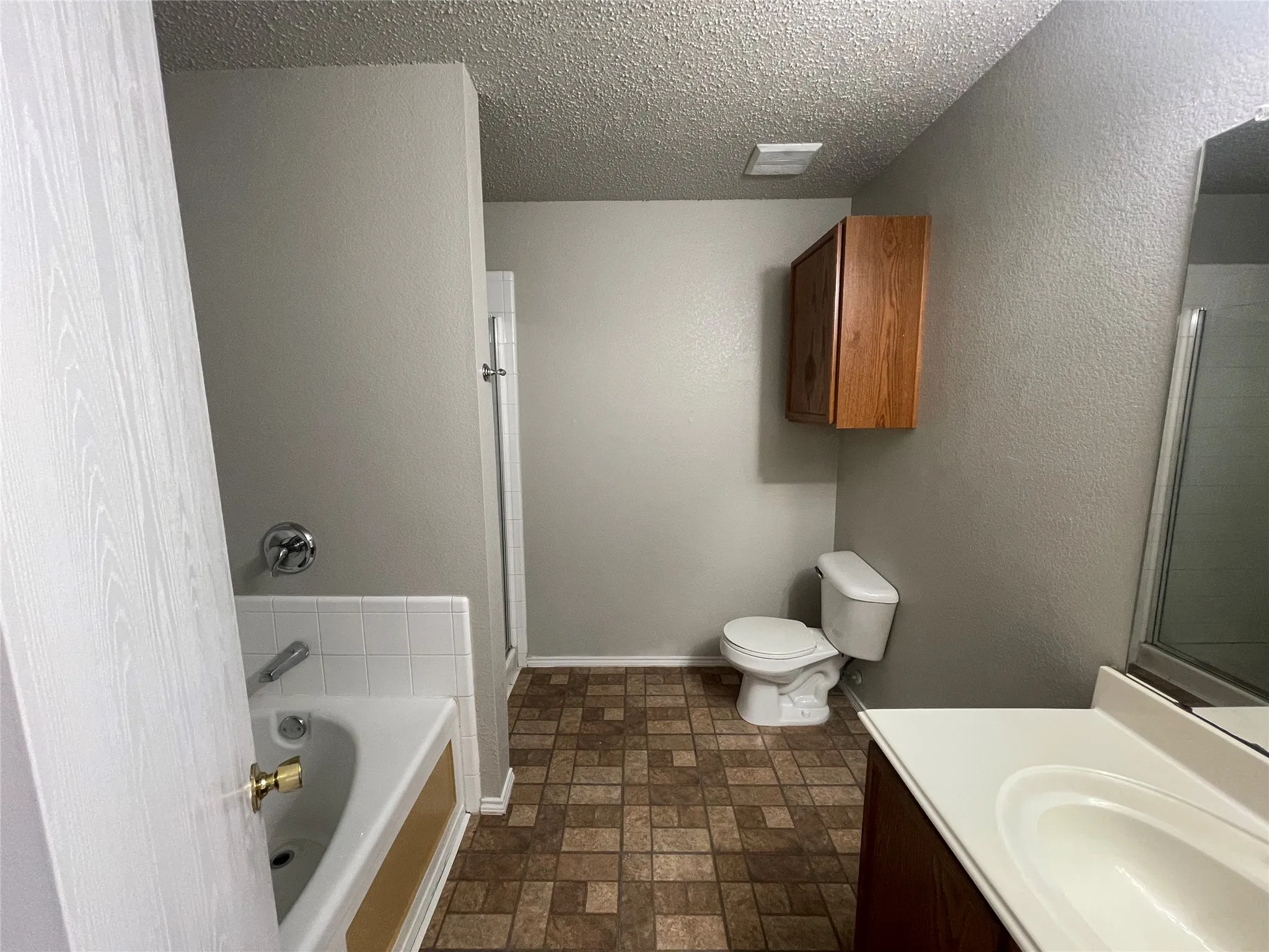 Bathroom featuring a vanity with an integrated sink, a toilet, a bathtub with white tile surround, a shower enclosure, and a wall-mounted wooden cabinet