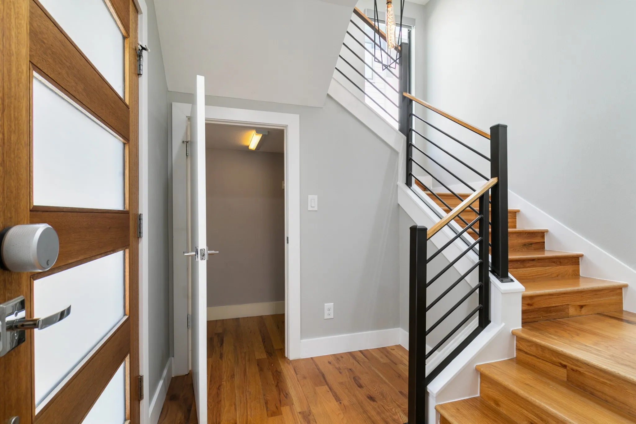 Wood-finish flooring, modern wood-panel door with frosted glass, open doorway with interior lighting, wood stairs, and contemporary metal railing with wood handrail
