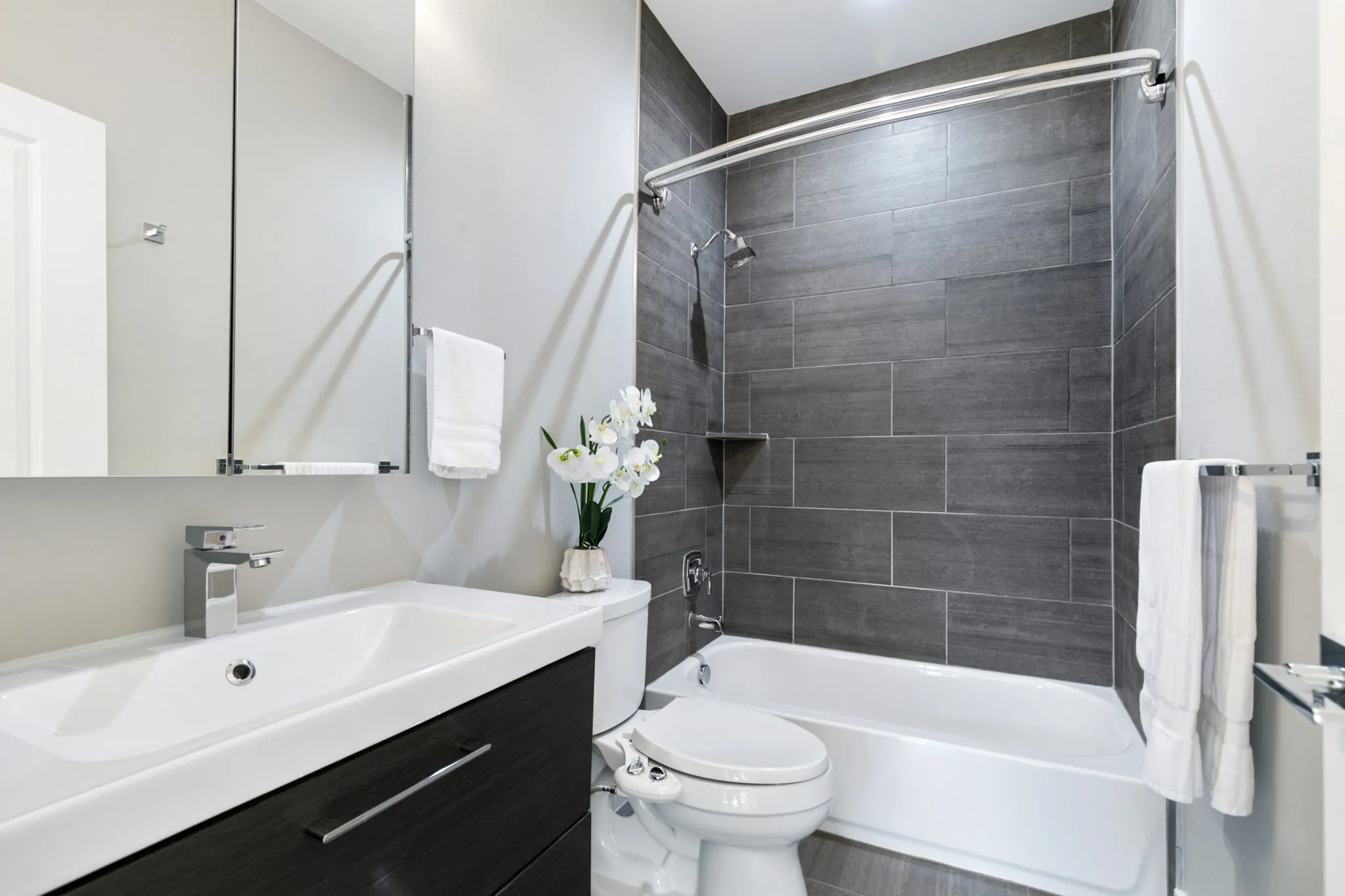 Vanity with integrated sink, chrome faucet, medicine cabinet, bathtub with dark tile surround, and wall-mounted showerhead