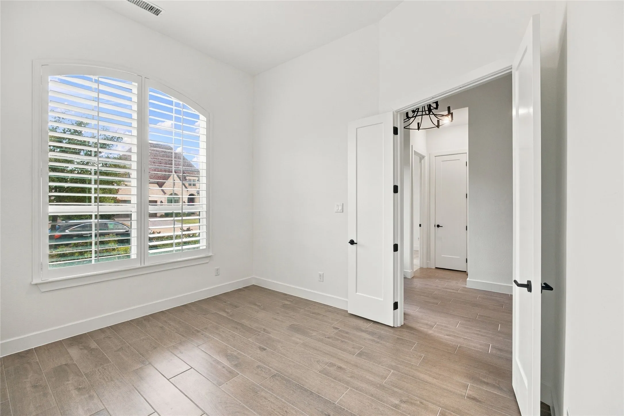 Arched window with plantation shutters, wood-finish tile flooring, double doors with matte black hardware, and crisp white walls
