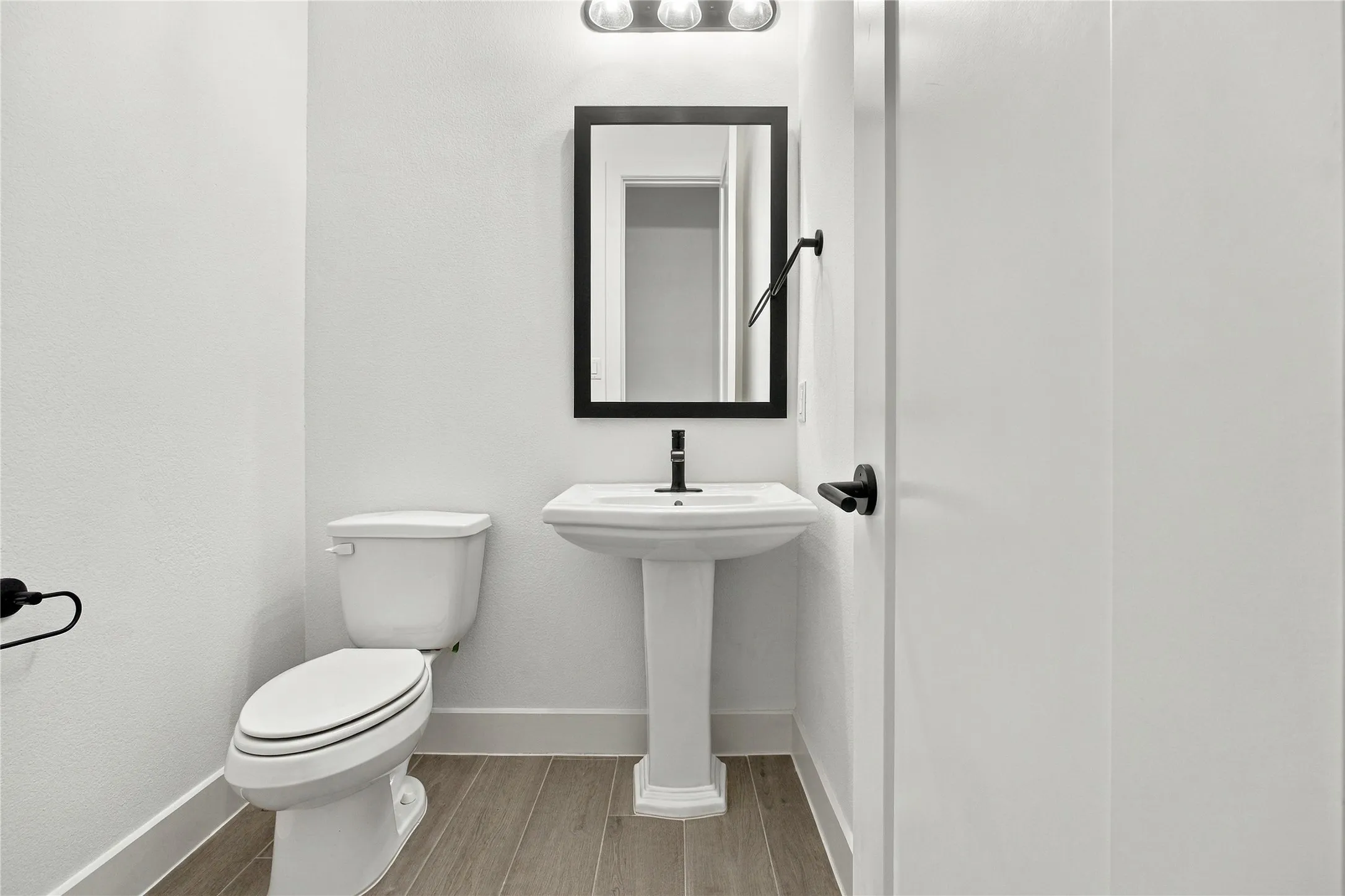 Hallway half-bath features pedestal sink with a rectangular mirror, wood-finish flooring