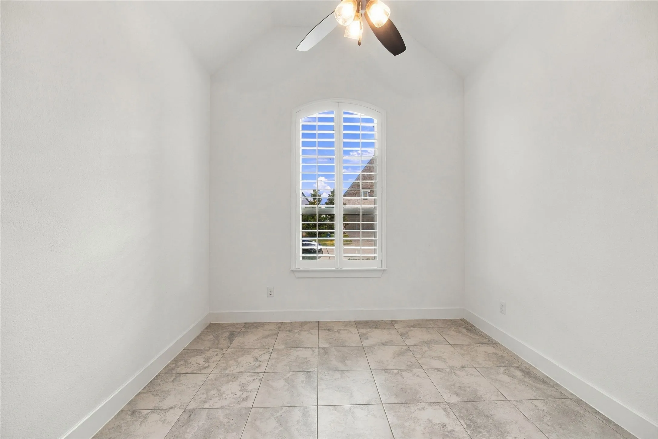 Vaulted ceiling with an attached ceiling fan, arched window with plantation shutters, and large format tile flooring, ensuite bath