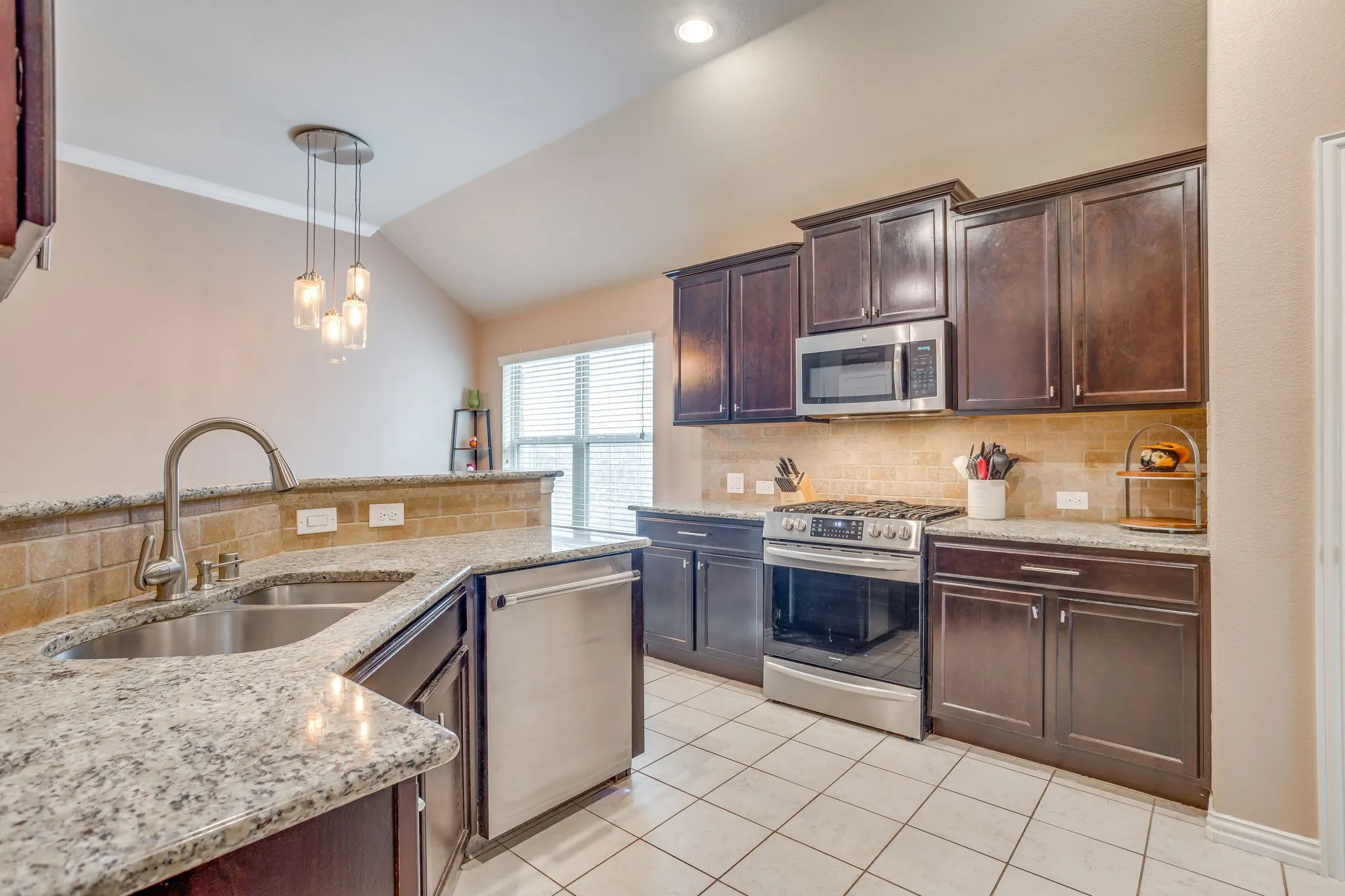 Kitchen featuring dark wood cabinetry, granite countertops, a tile backsplash, stainless steel appliances, and a double basin sink