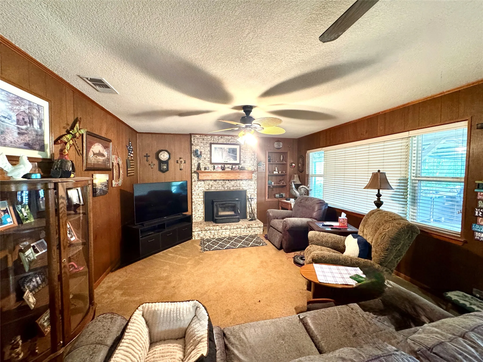 Brick-faced fireplace with a hearth insert, wood paneling, built-in shelving, ceiling fan, and carpeted flooring