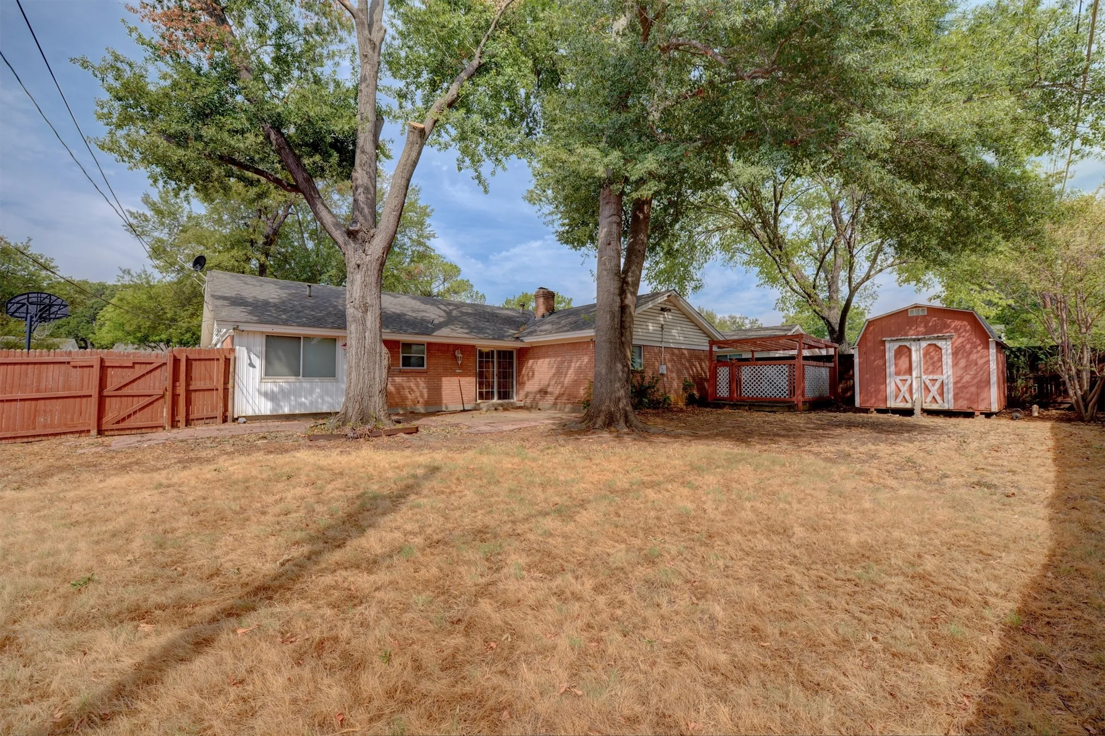 Expansive backyard featuring mature trees, a red wood fence with gate, and a storage shed with white trim