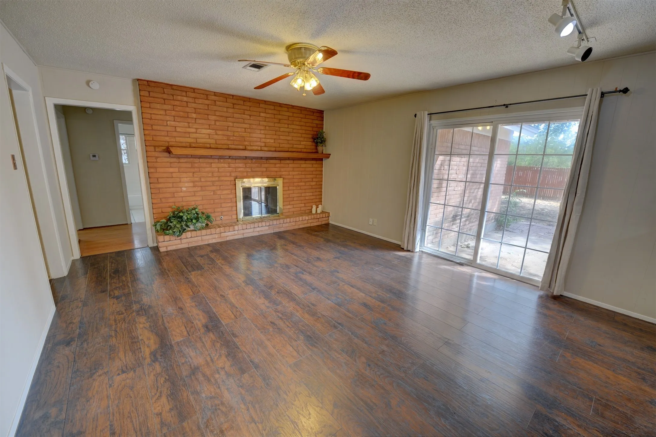 Brick-faced fireplace with hearth and mantel, wood-finish flooring, sliding glass door, track lighting, and a ceiling fan