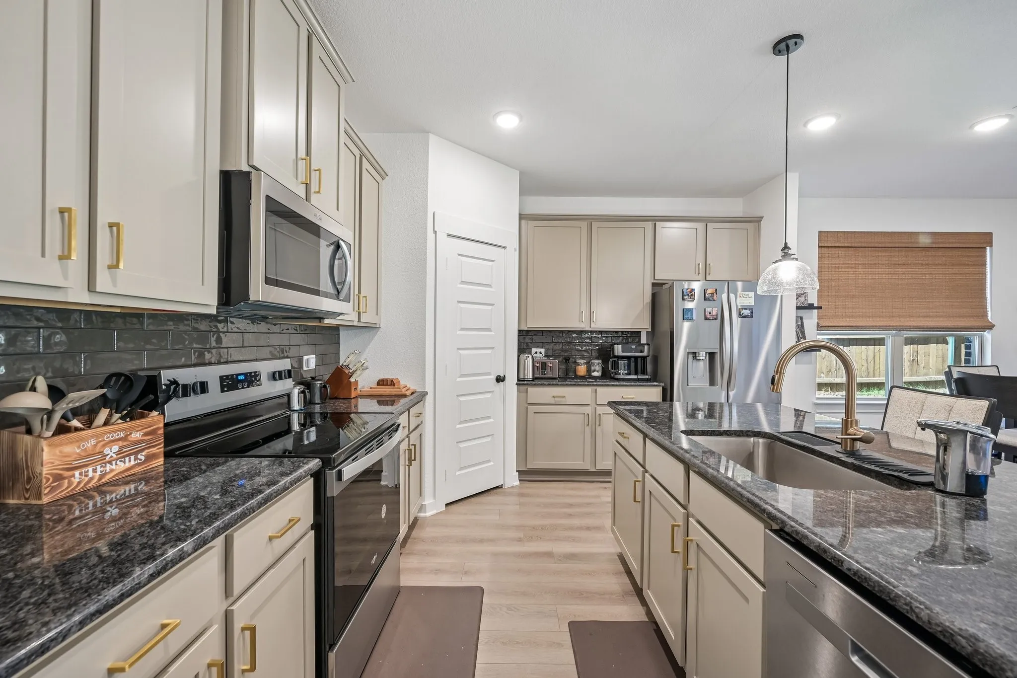 Kitchen featuring light-toned cabinetry with gold-finish hardware, dark subway tile backsplash, stainless steel appliances, a center island with an undermount sink, and wood-finish flooring