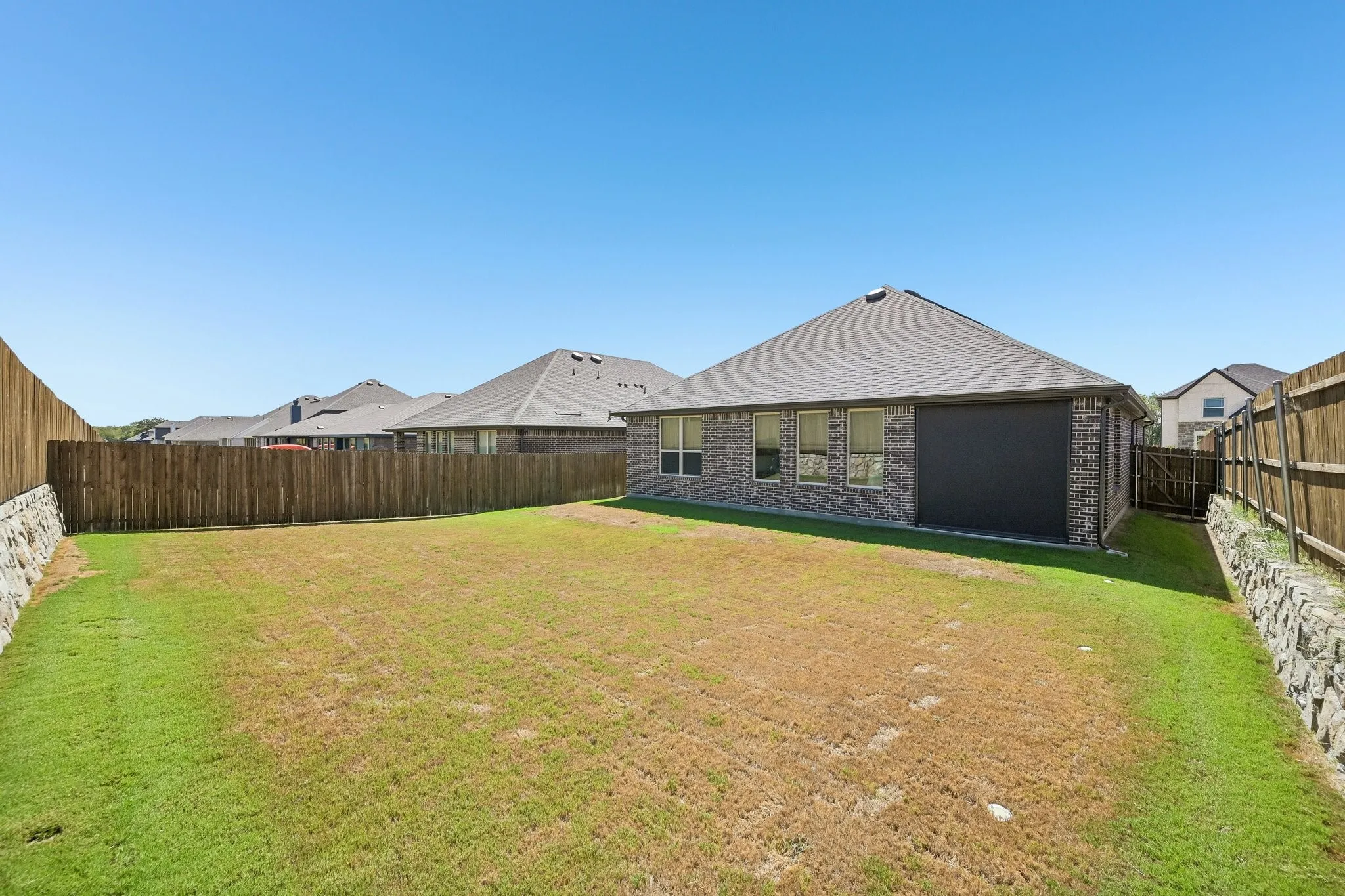 Spacious backyard featuring a wood privacy fence, stone retaining wall, and a brick exterior home with a shingle roof