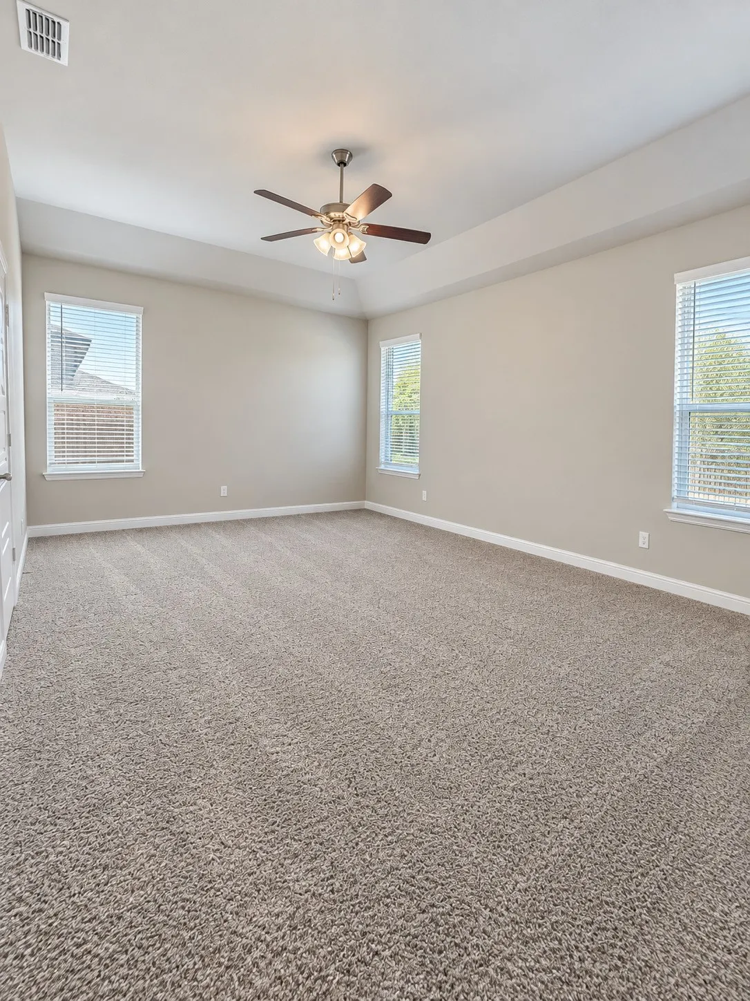 Spacious room featuring neutral carpet flooring, multiple windows with blinds, a tray ceiling, and a ceiling fan with integrated lighting