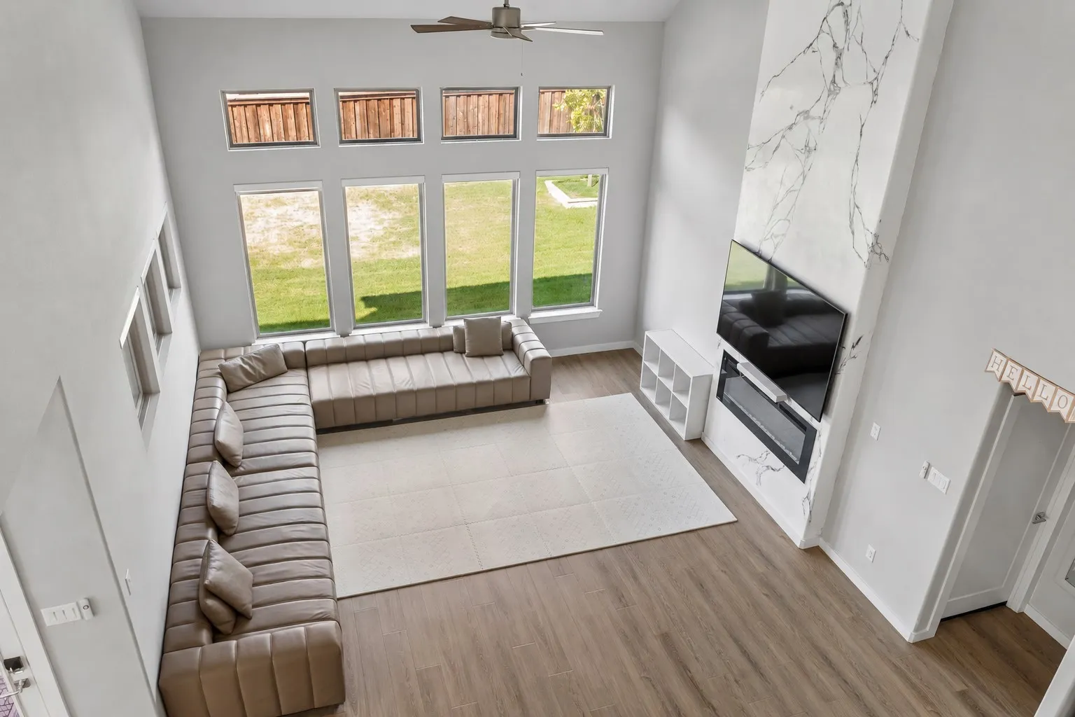 Two-story living area with high ceilings, multiple large windows, wood-finish flooring, a ceiling fan, and a fireplace with a white and gray veined surround