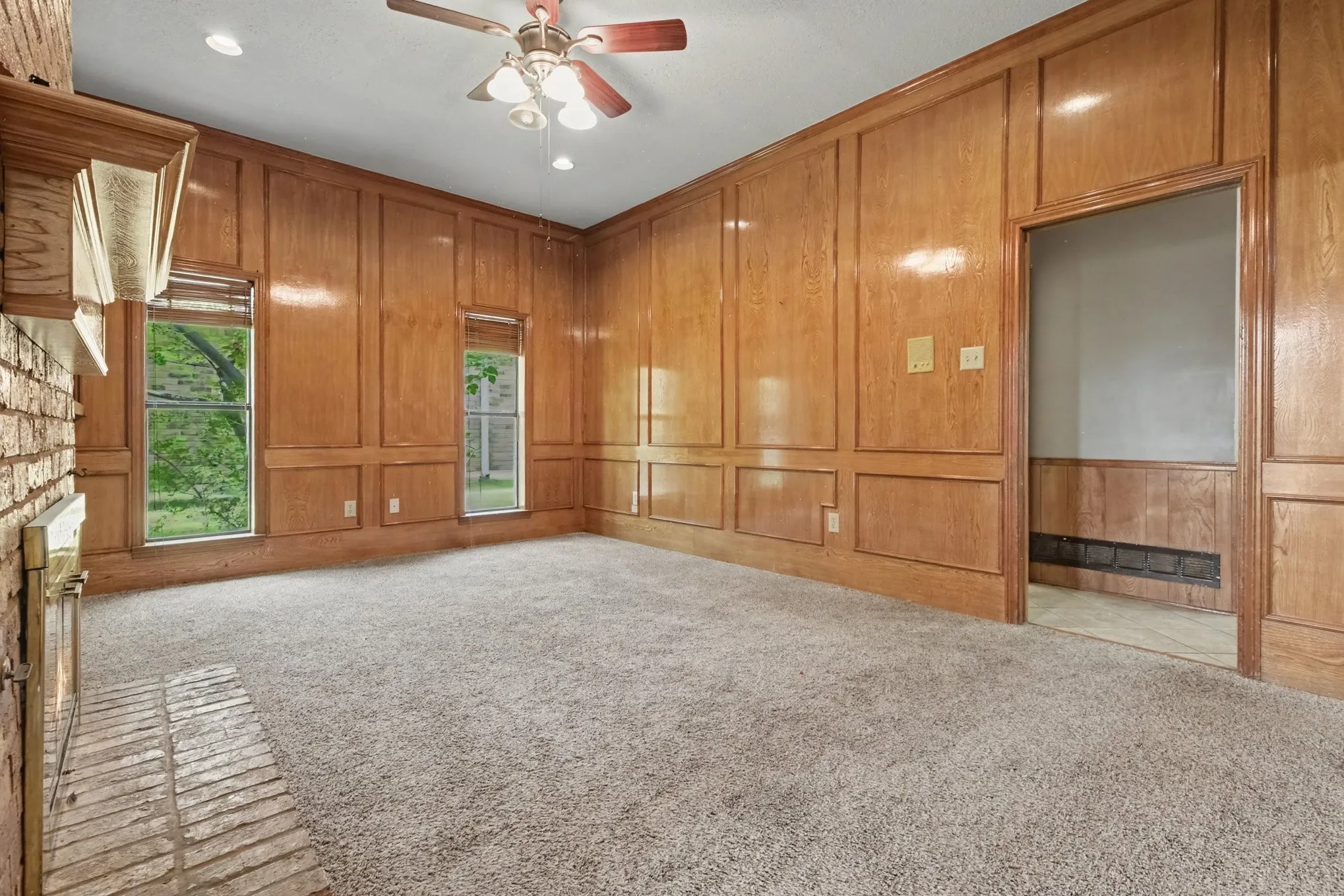 Wood paneling with decorative trim, brick fireplace with mantel, ceiling fan with integrated lighting, and carpeted flooring