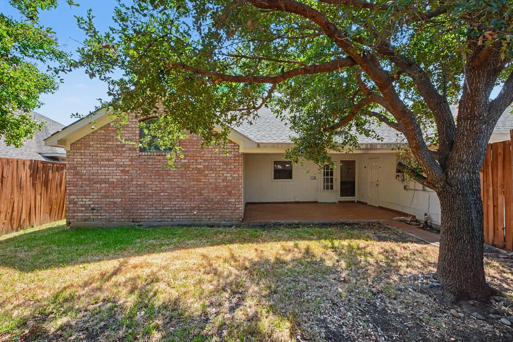 Expansive backyard featuring a mature tree, brick exterior, wood fence, and a covered patio