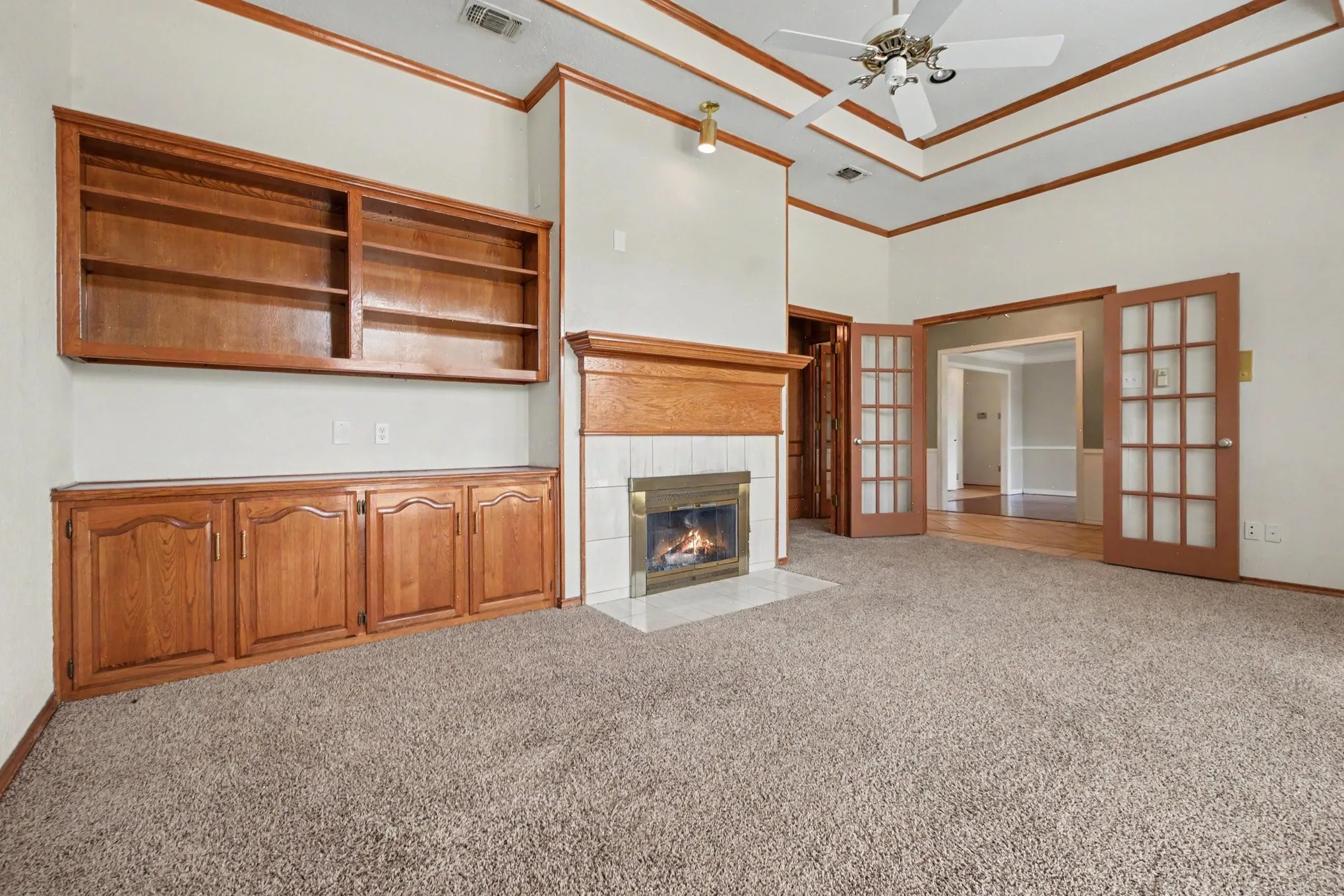 Built-in wood cabinetry and shelving, a fireplace with a tile surround and wood mantel, a coffered ceiling with a ceiling fan, and French doors