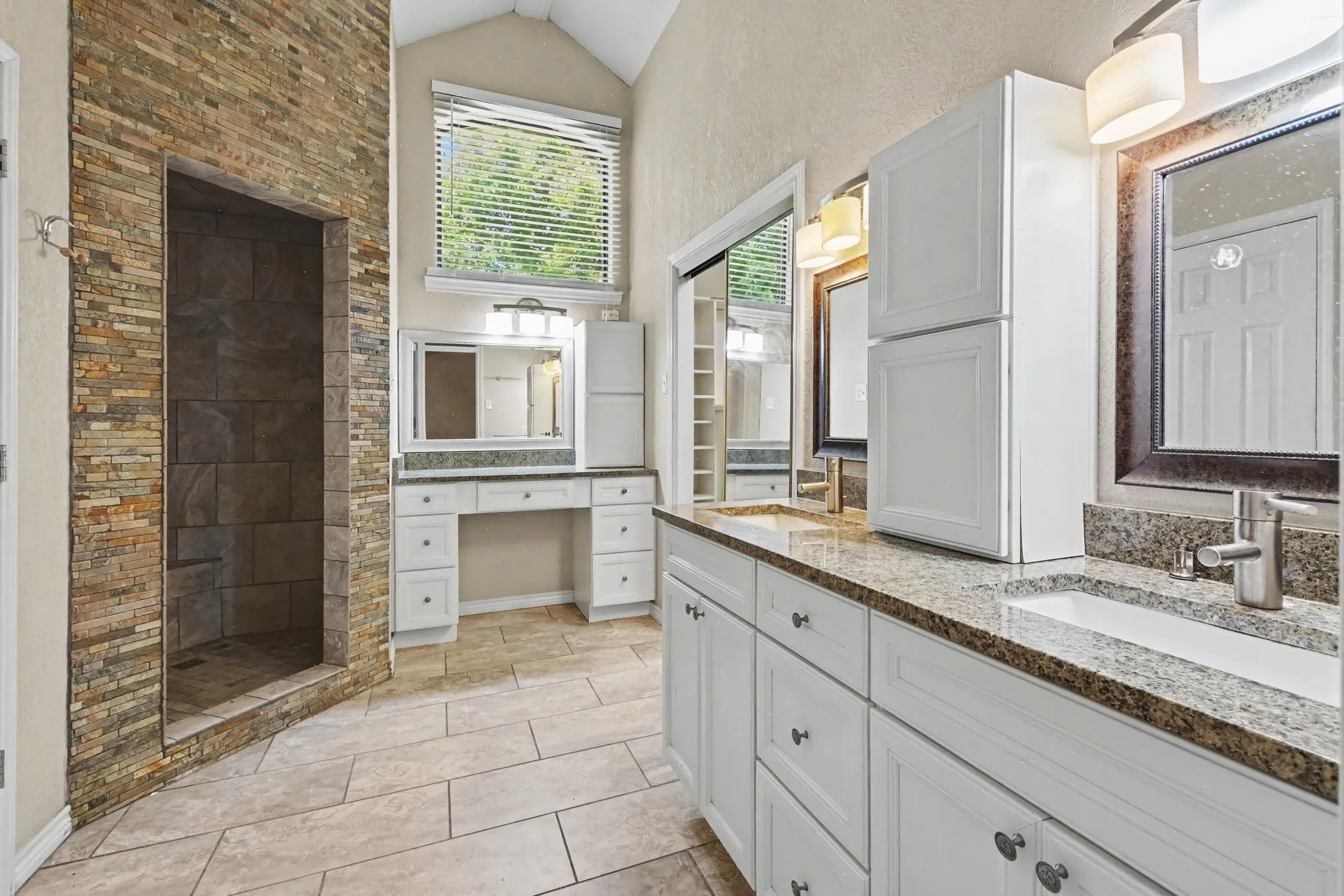 Spacious bathroom featuring a stone-tiled walk-in shower, dual vanities with granite countertops, white cabinetry, and tiled flooring