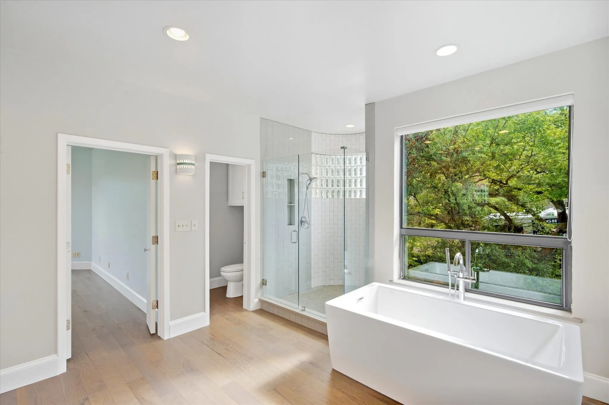 Freestanding soaking tub with a floor-mounted faucet, glass-enclosed shower with subway tile, wood-finish flooring, and recessed lighting