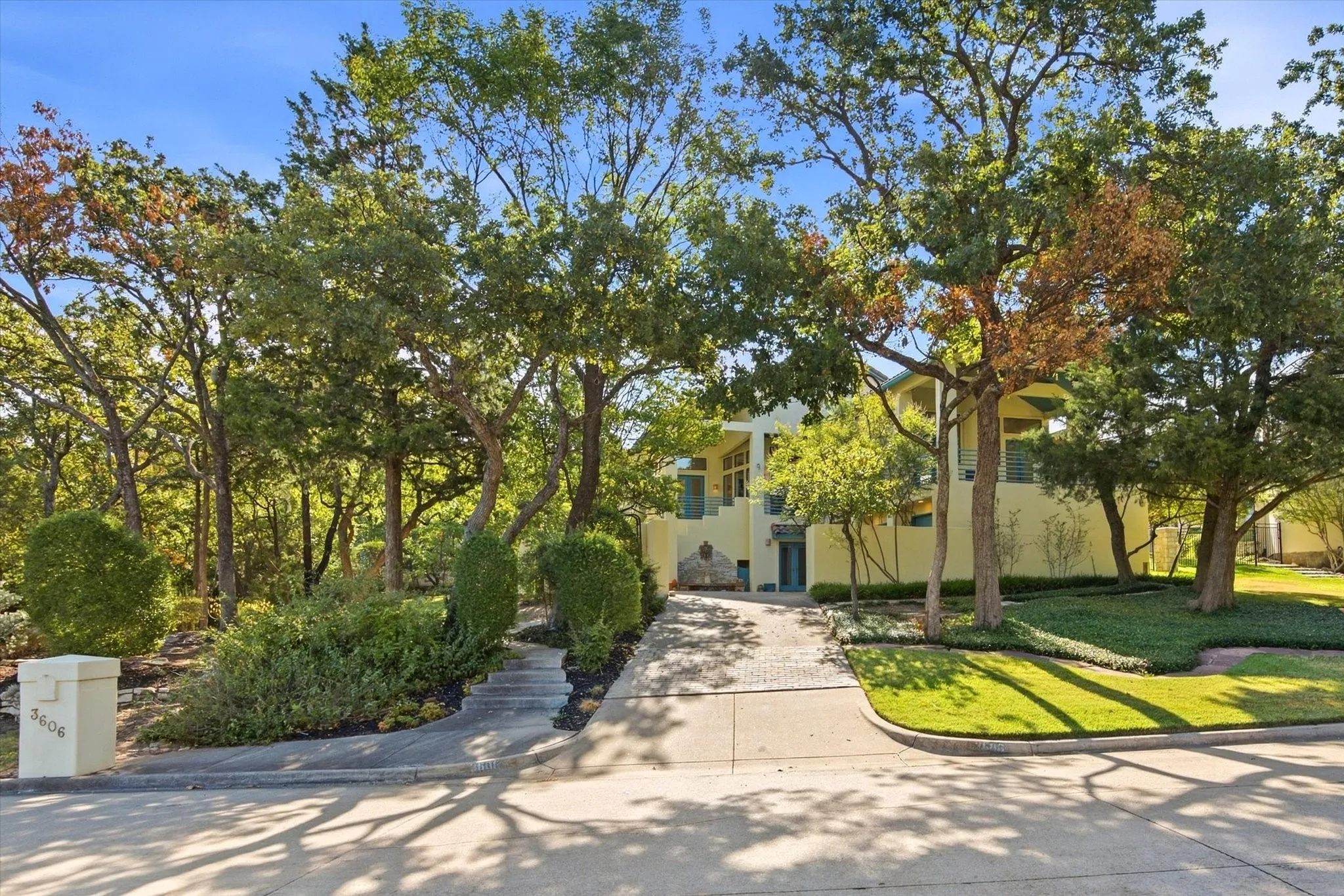 Yellow stucco exterior, mature trees, landscaped grounds, concrete driveway, and stone staircase