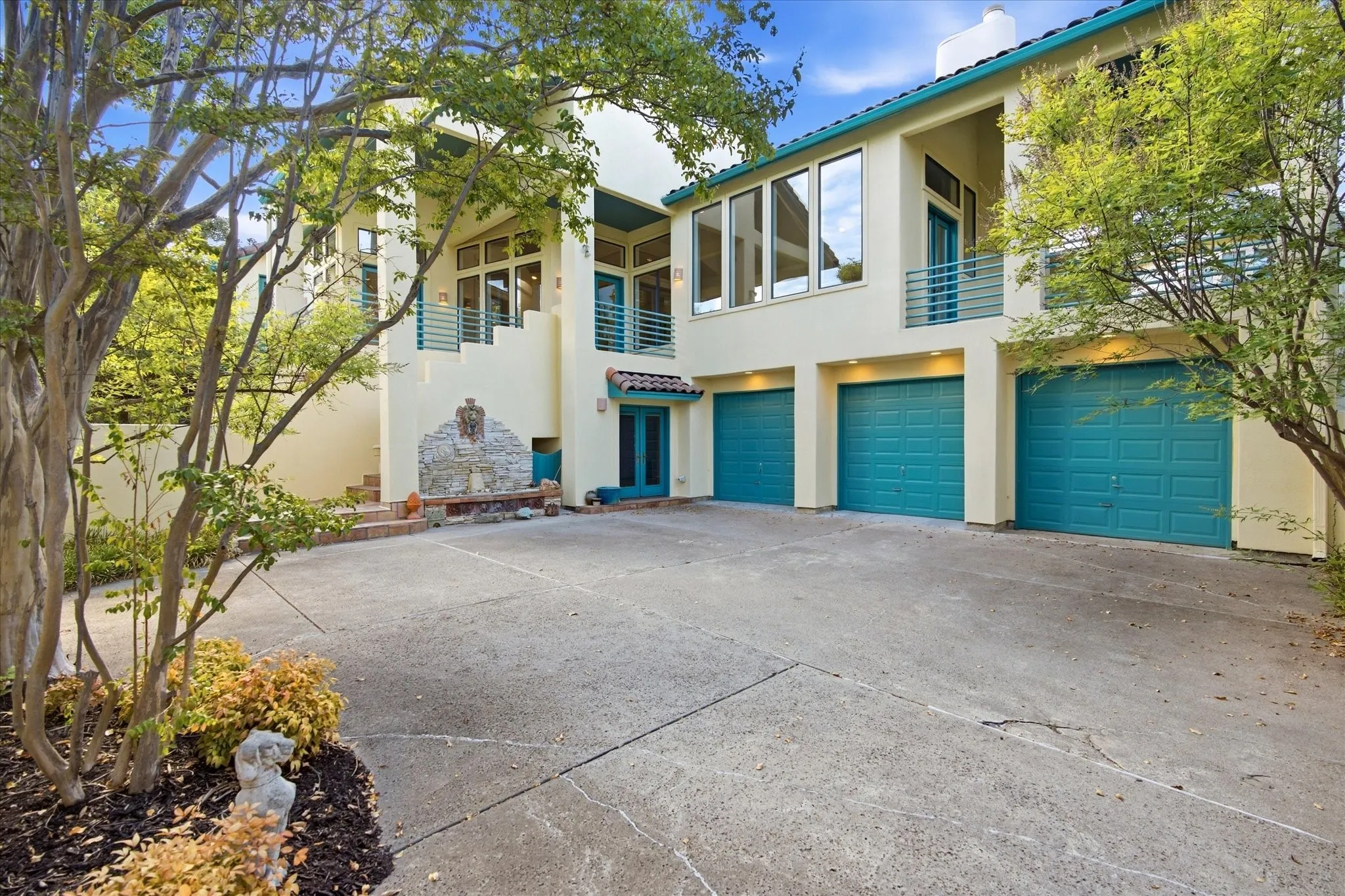 Stucco exterior with multiple balconies, three garage doors, a decorative water feature, and mature trees