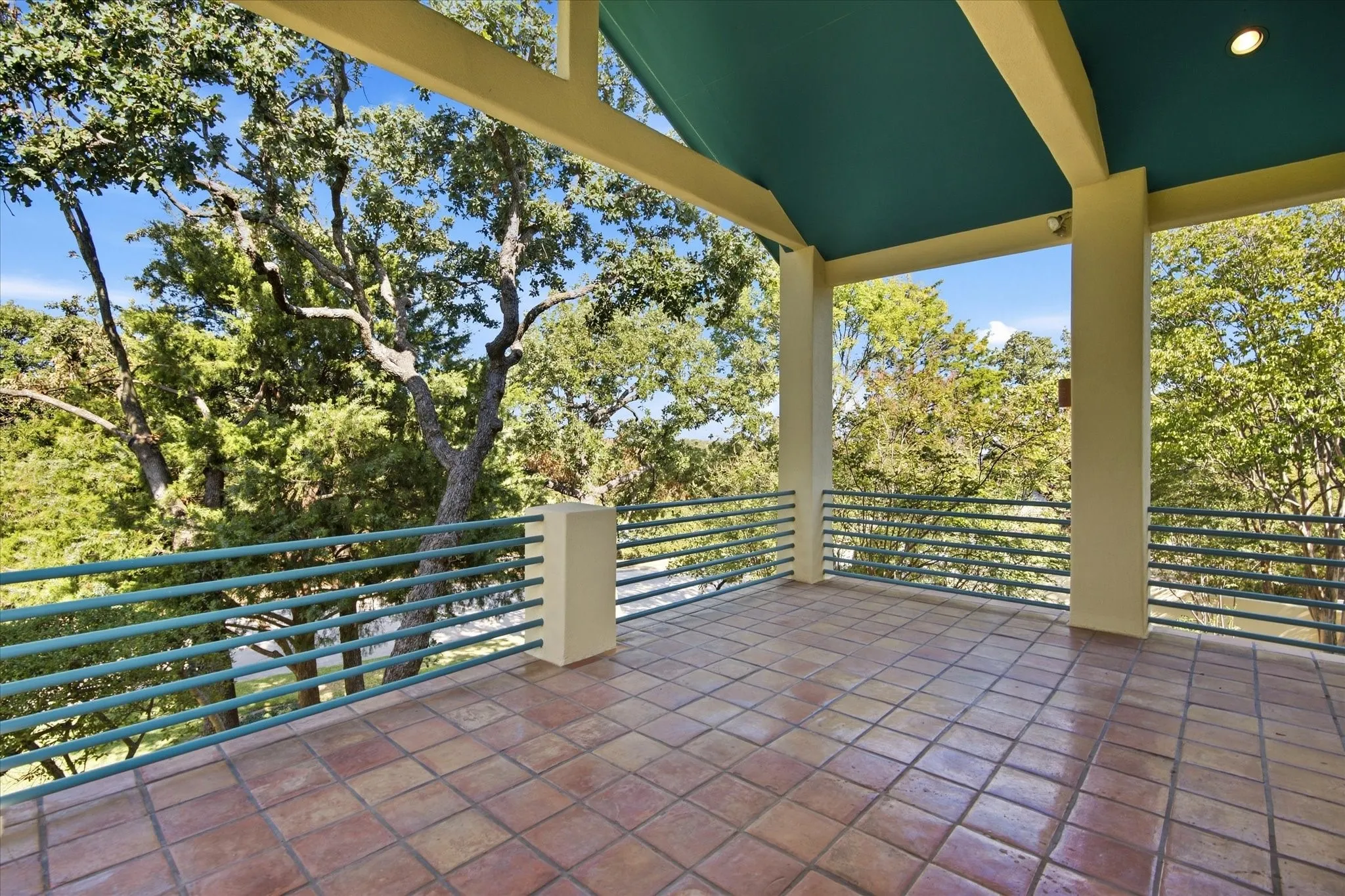 Expansive outdoor balcony featuring terracotta tile flooring, a sloped ceiling with recessed lighting, sturdy support columns, and an extensive metal railing