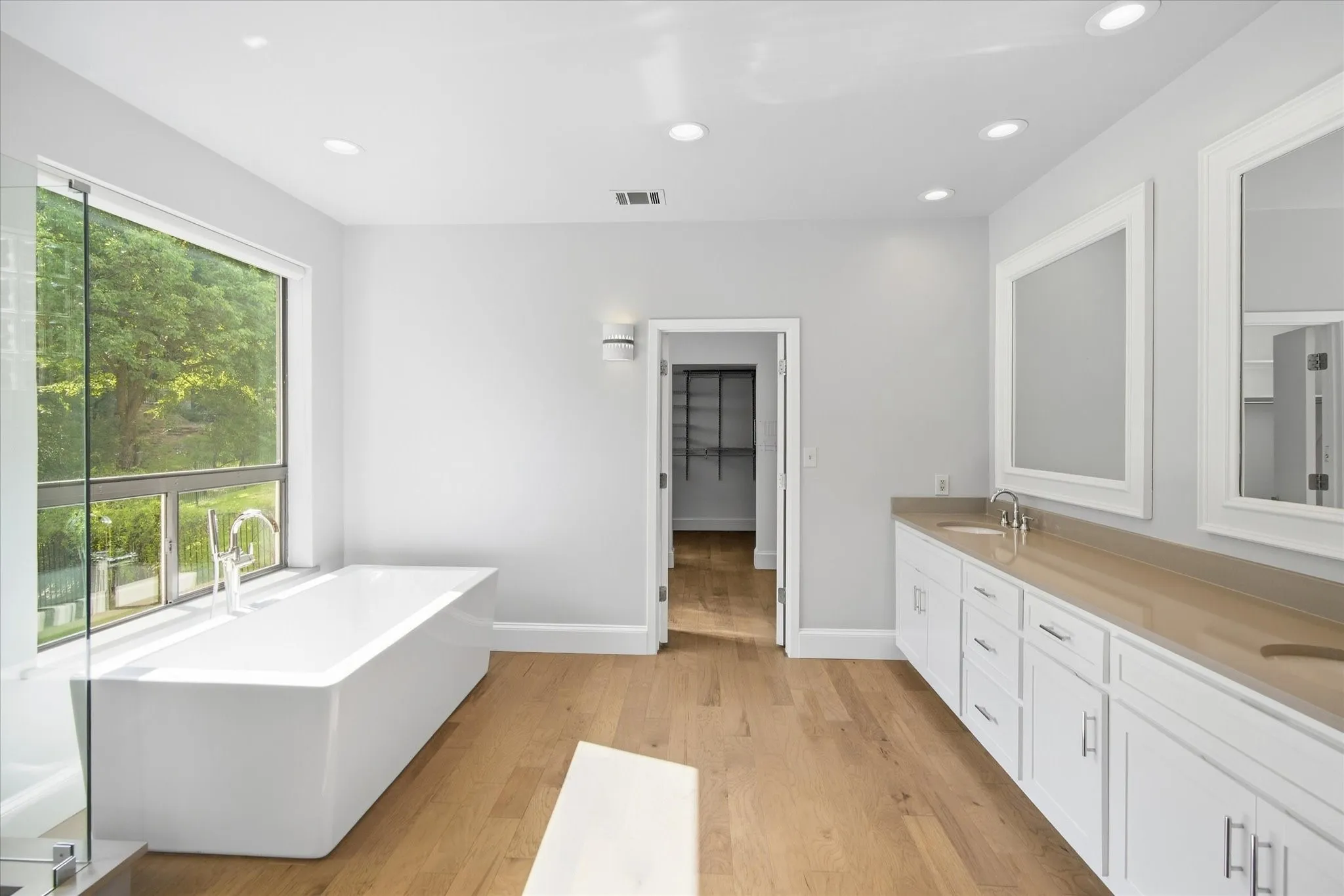 Freestanding soaking tub with a full-height window, dual vanity with undermount sinks, wood-finish flooring, and recessed lighting