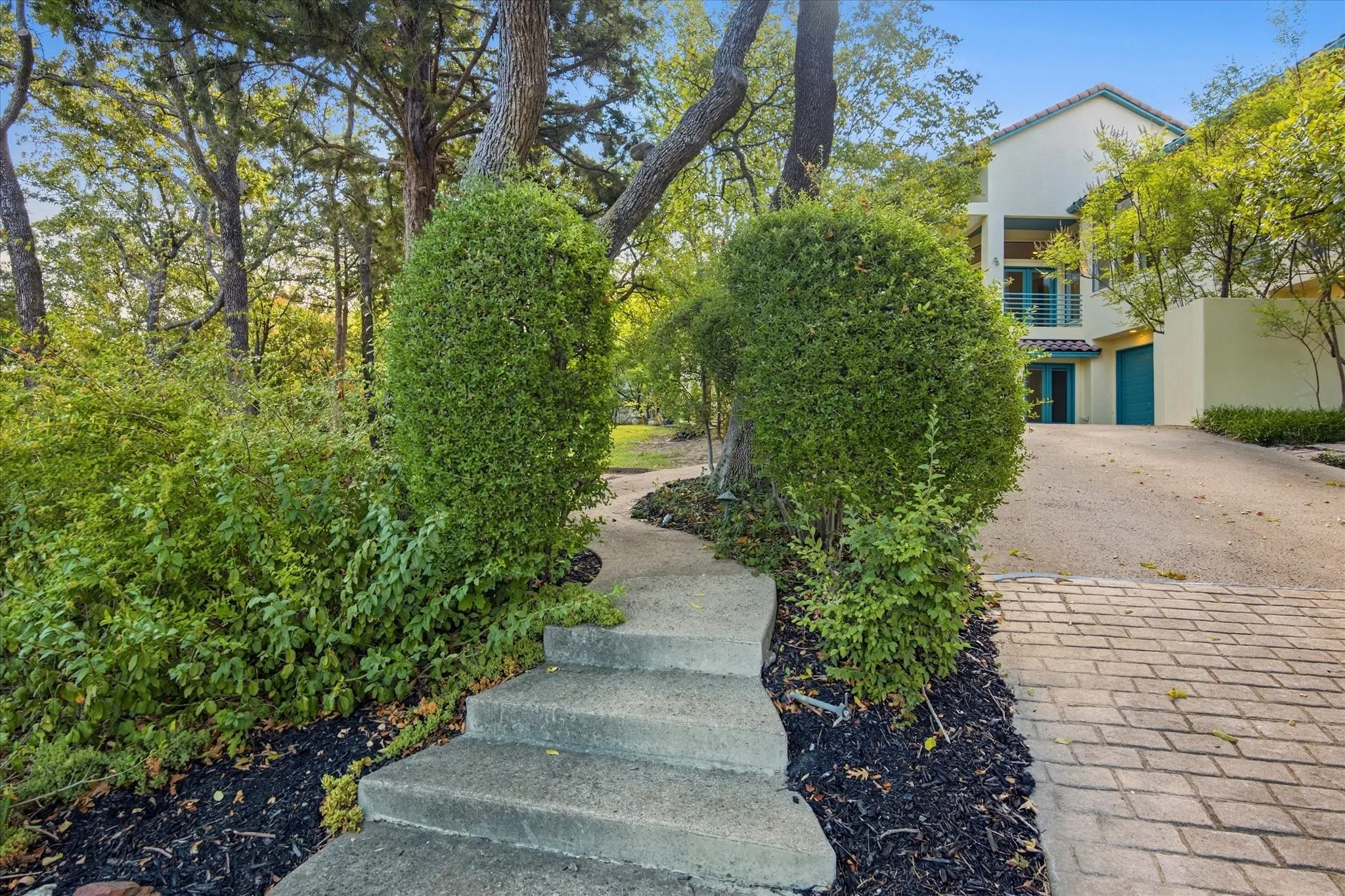Curved concrete pathway with steps and mature landscaping, leading to a light-colored stucco exterior with a tiled roof