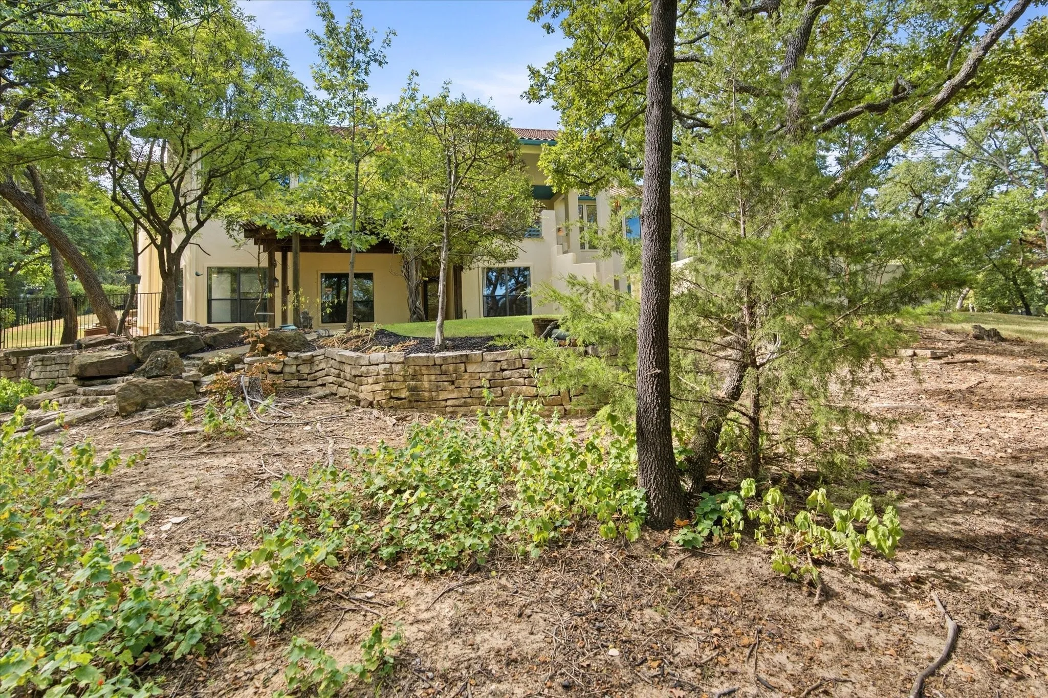 Stucco exterior with multiple windows, covered patio, and natural landscaping with mature trees and stone retaining walls