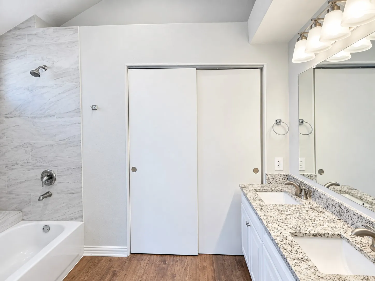 Bathroom featuring a dual vanity with a granite-style countertop, undermount sinks, wood-finish flooring, a bathtub with a shower, and sliding closet doors