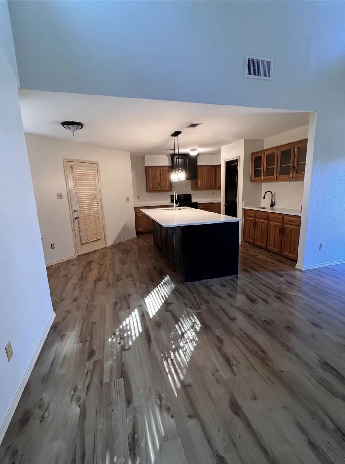 Wood-finish flooring extending through the space, kitchen island with a light-colored countertop, overhead pendant lighting, and abundant wood cabinetry with glass-front accents