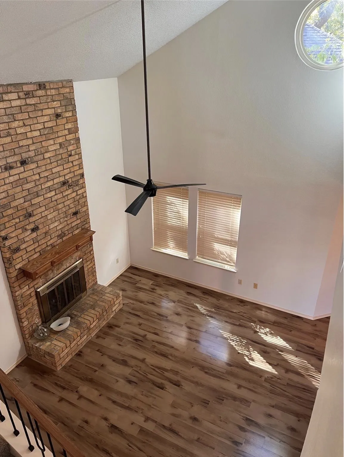 Vaulted ceiling with a black ceiling fan, brick fireplace, wood-finish flooring, and two windows
