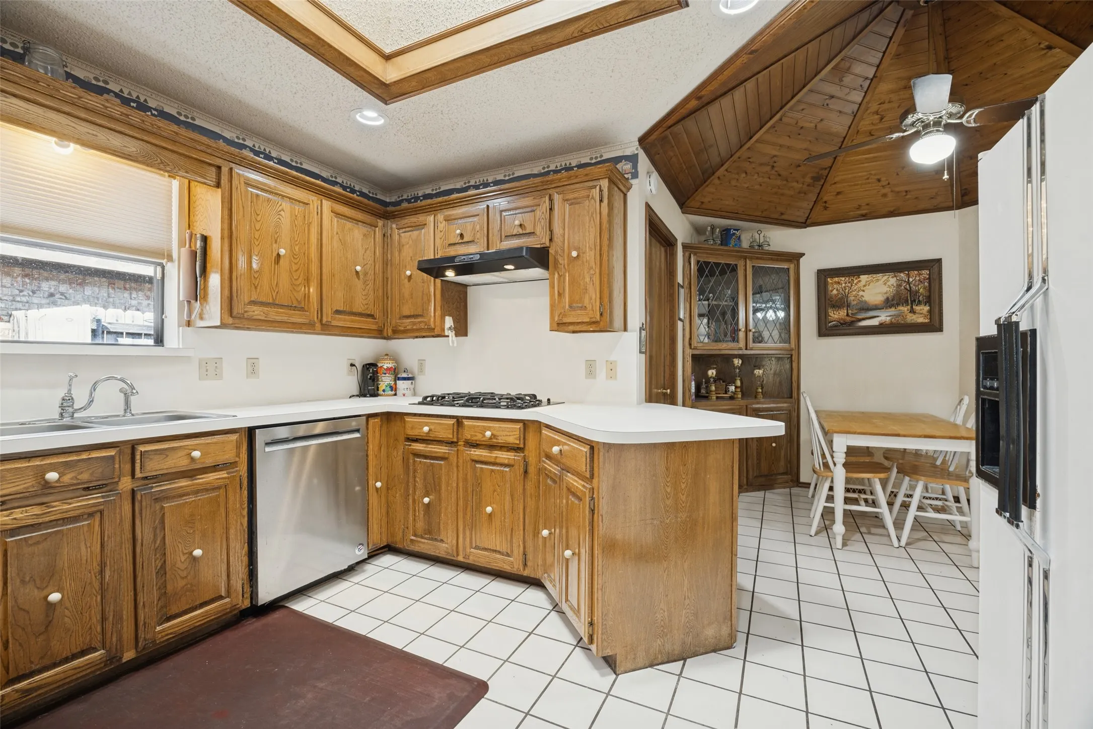 Oak cabinetry with stainless steel dishwasher, gas cooktop with exhaust hood, and white tile flooring