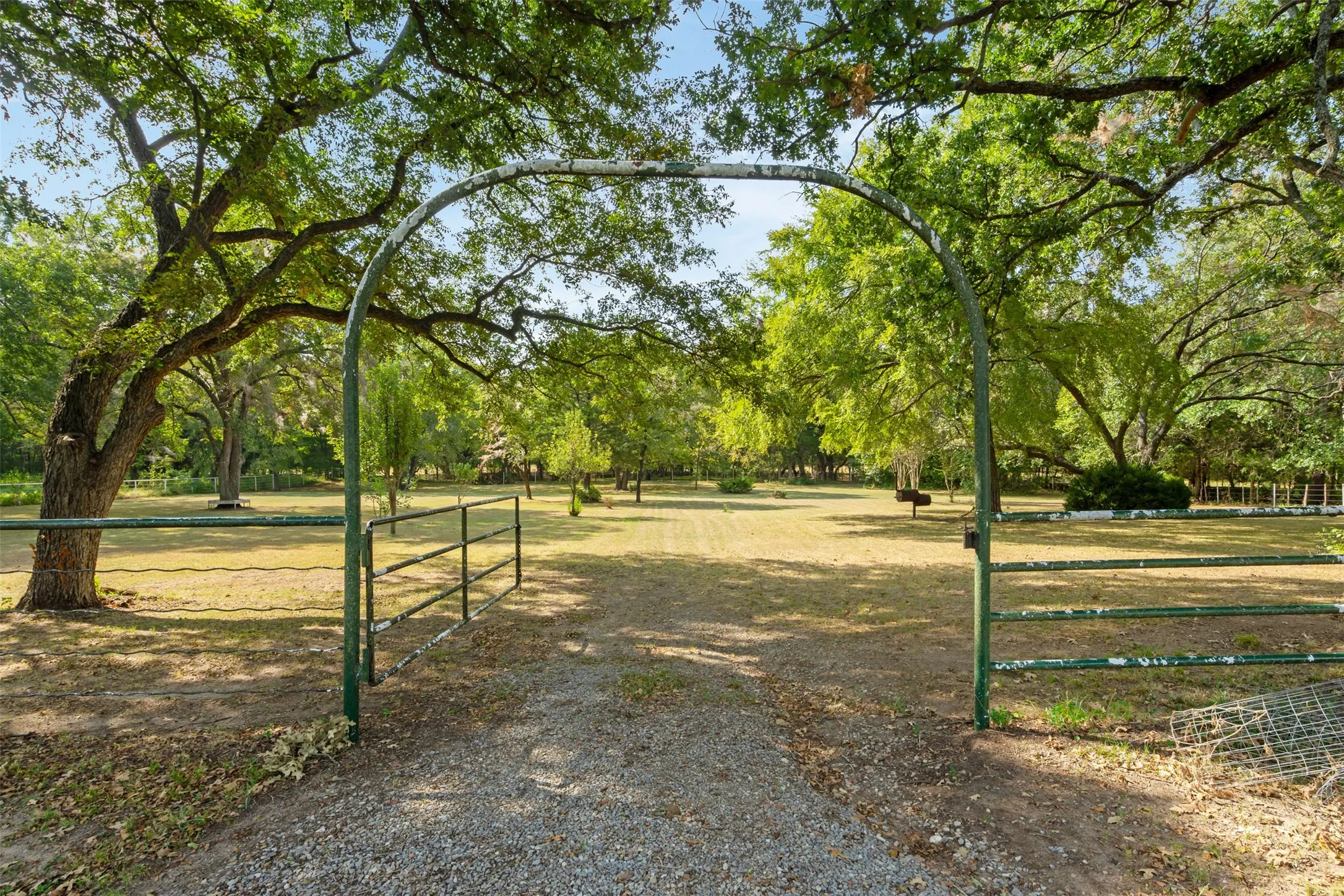 Gravel drive entering through a metal archway and gate, bordered by mature trees and expansive grassy areas