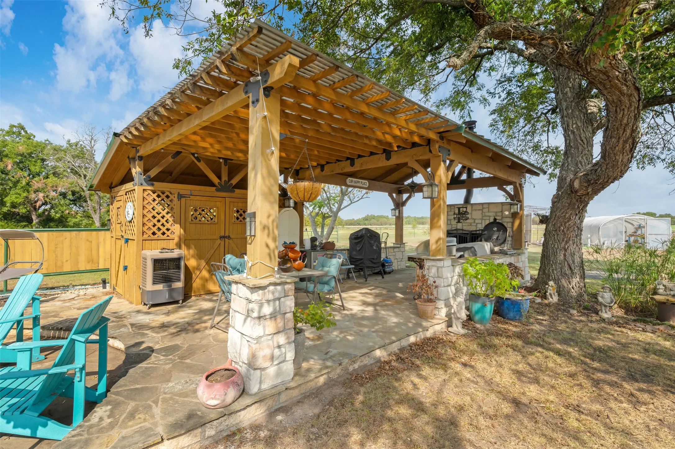 Expansive outdoor entertaining area featuring a stone patio, a substantial wooden pergola with a corrugated roof, a built-in stone outdoor kitchen, and a separate lattice-accented storage structure
