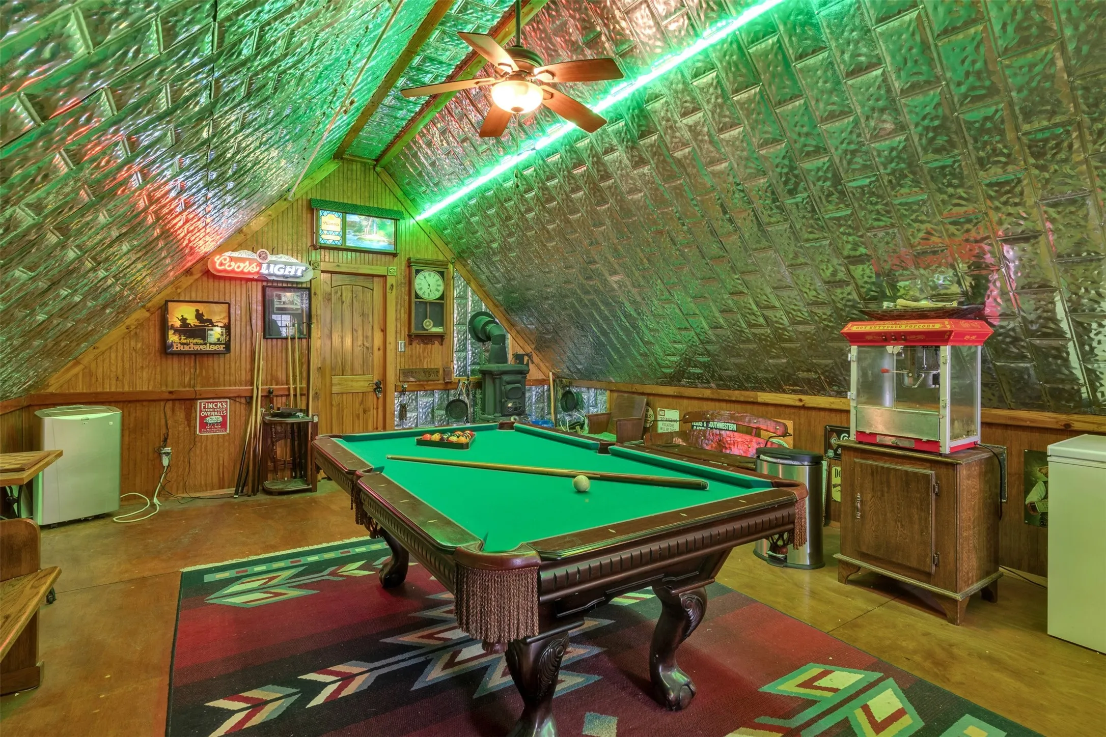 Vaulted ceiling with textured metallic panels, wood-paneled walls, ceiling fan, and built-in wood-burning stove