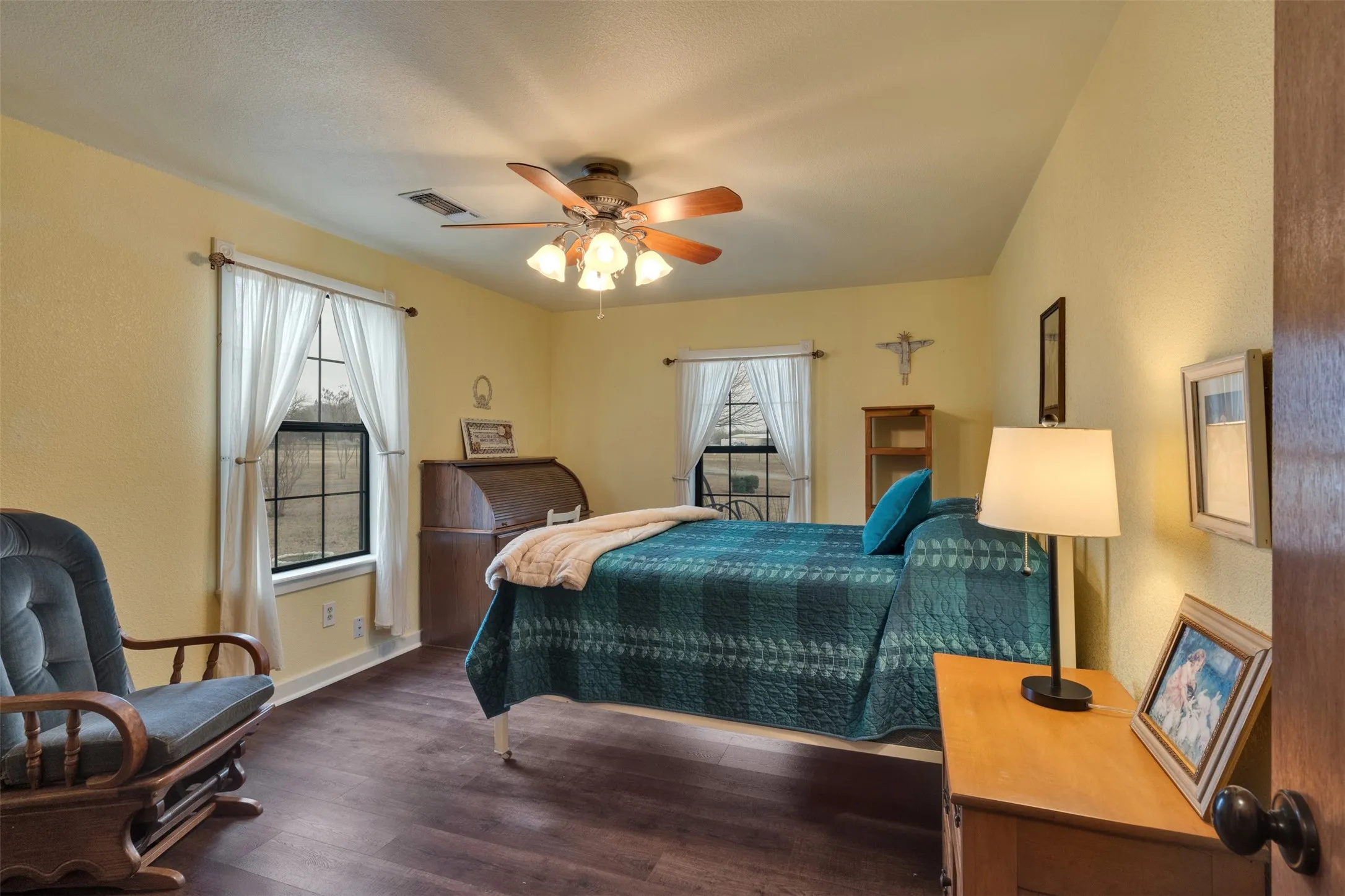 Wood-finish flooring, a ceiling fan with integrated lighting, and multiple windows with white curtains