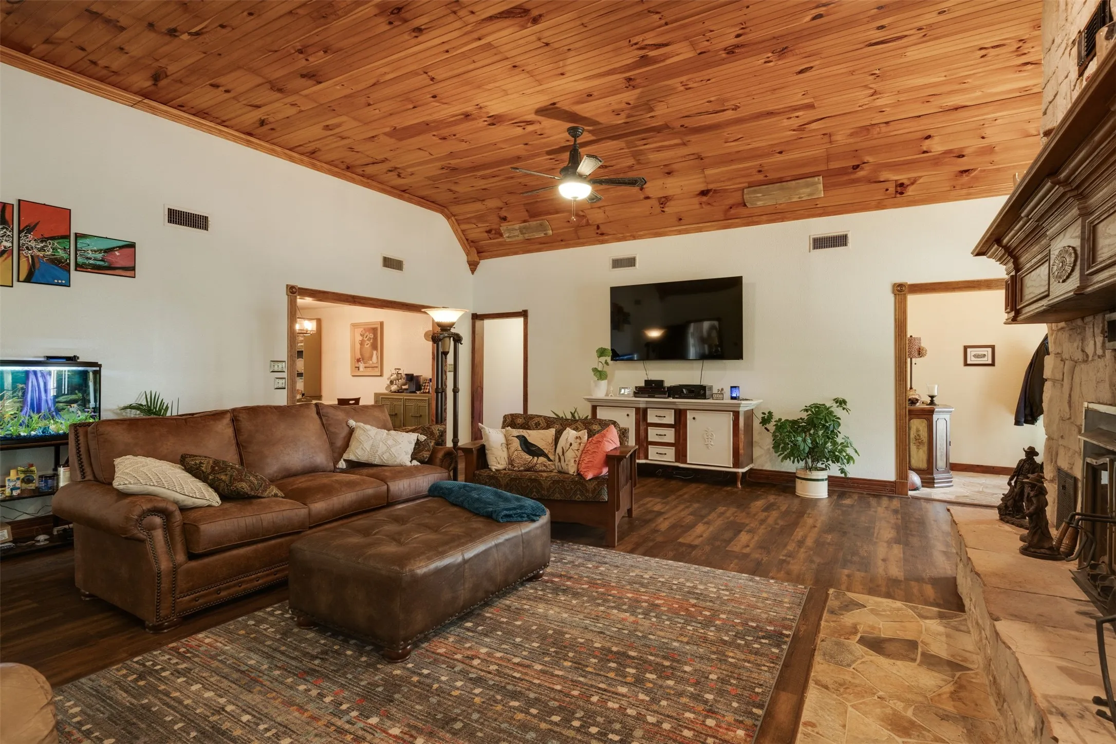 Vaulted wood-paneled ceiling with a central fan, stone fireplace with a decorative mantel, wood-finish flooring, and white walls