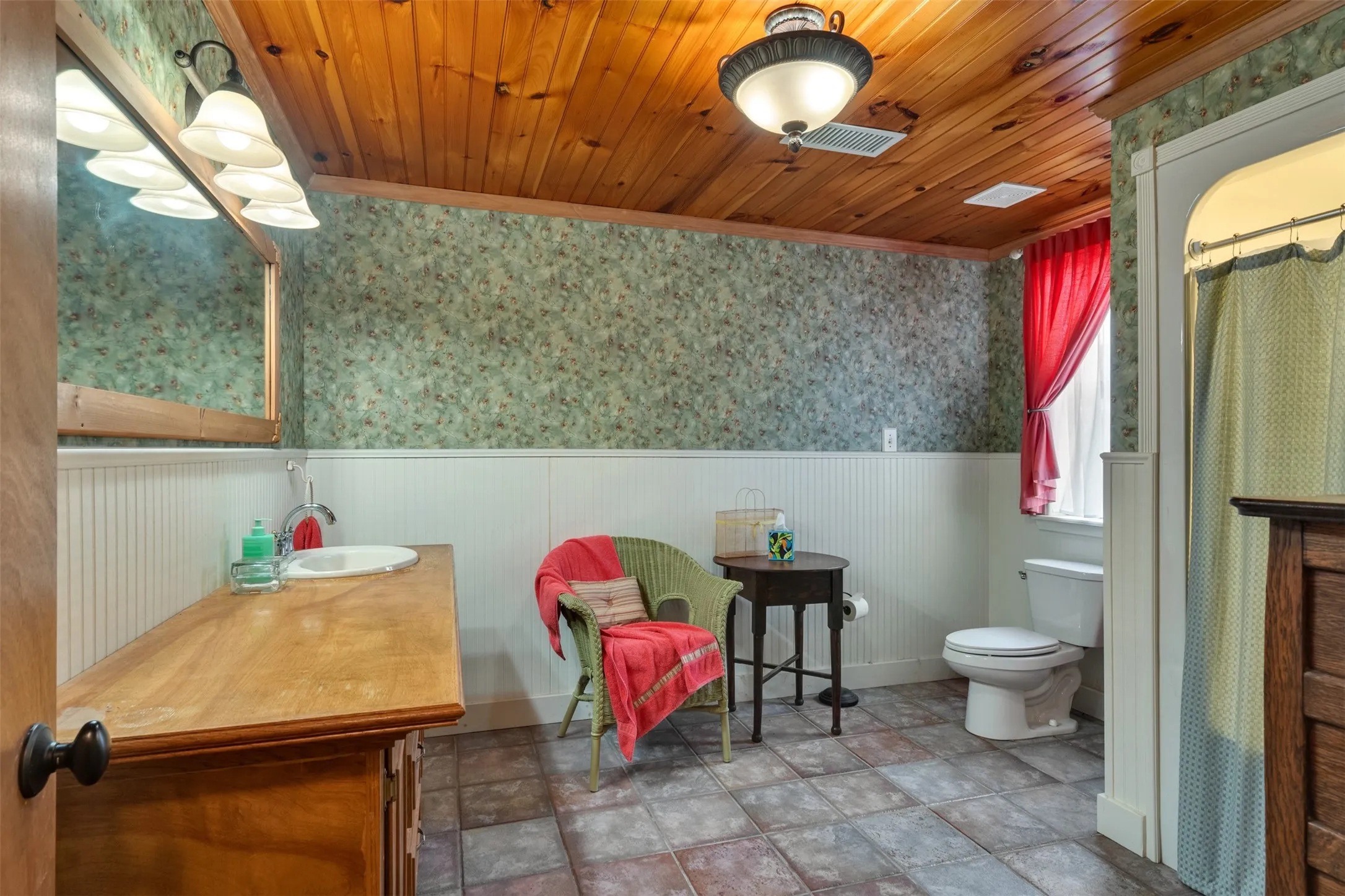 Wood plank ceiling, patterned wallpaper, white wainscoting, wood-finish vanity, and tiled flooring