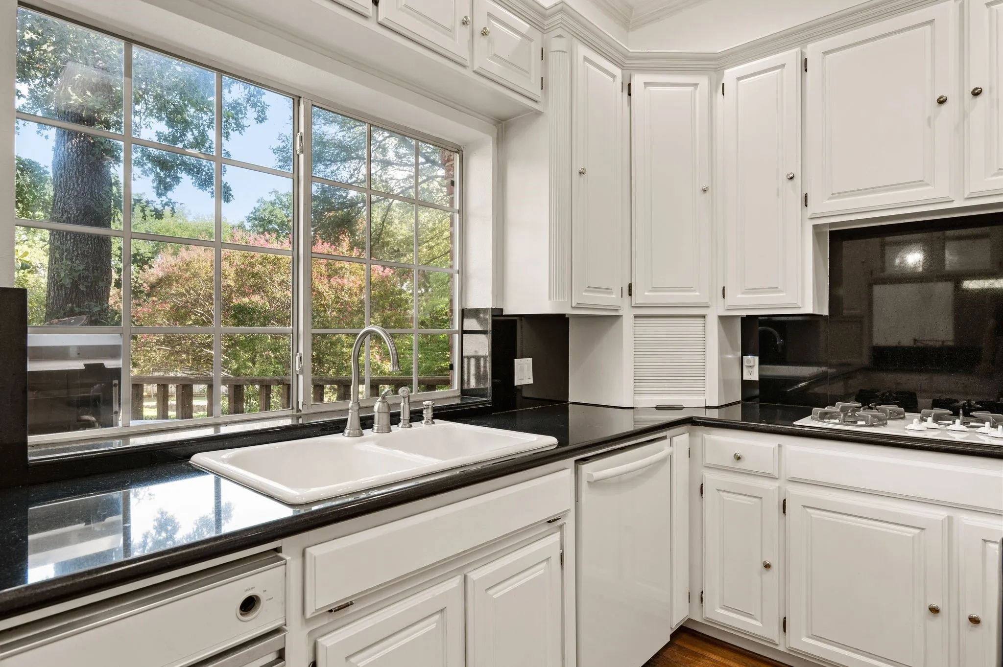 White cabinetry with brass-finish hardware, black countertops, dual-basin sink with stainless-finish faucet, and a large grid-pattern window