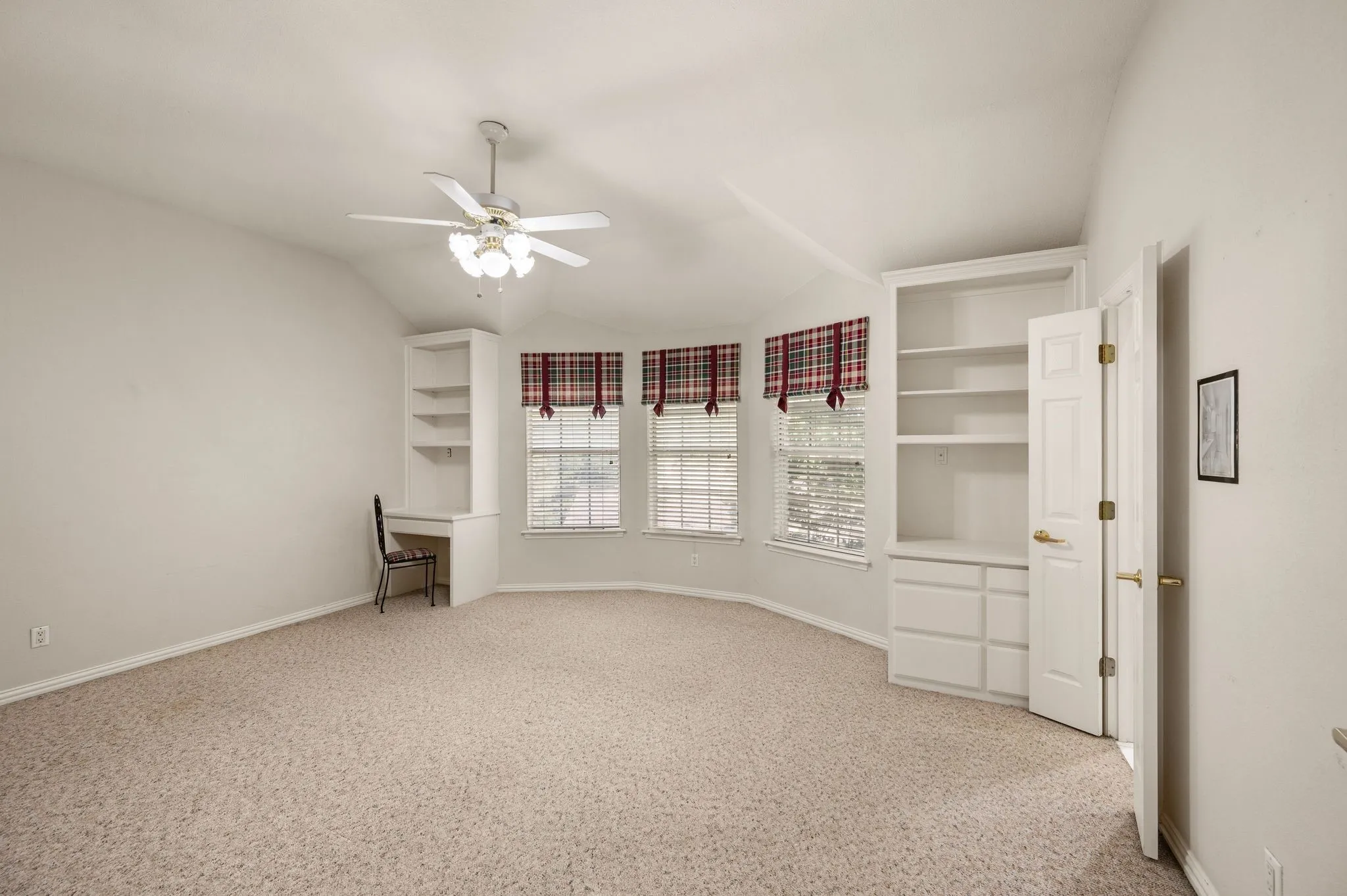 Tray ceiling with a ceiling fan, three windows with blinds, built-in shelving, and a built-in desk