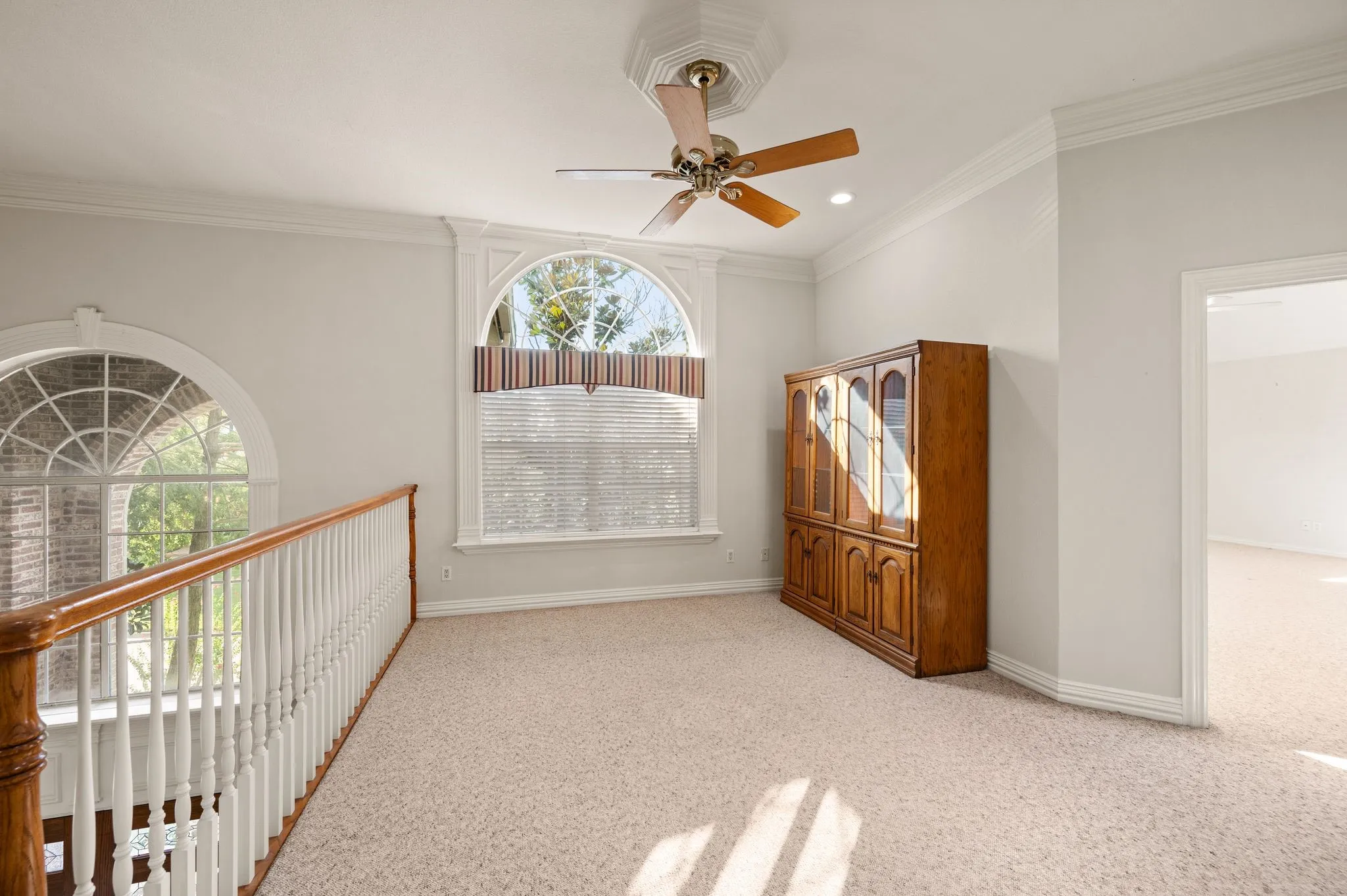 Carpeted loft area featuring a ceiling fan, arched windows, and crown molding