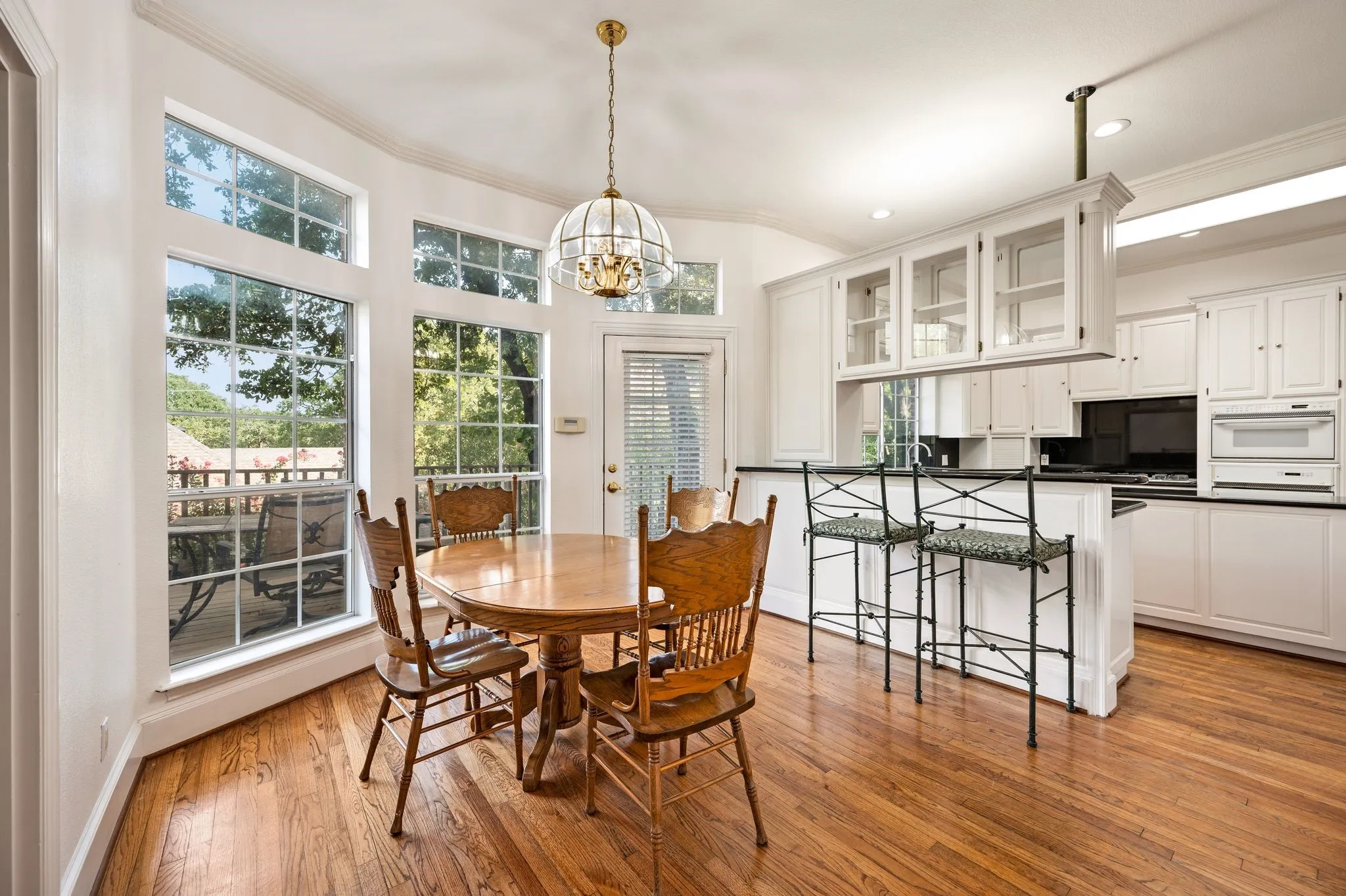 Hardwood flooring throughout, white cabinetry with glass-front uppers, built-in appliances, and a bay window dining area with a brass pendant light