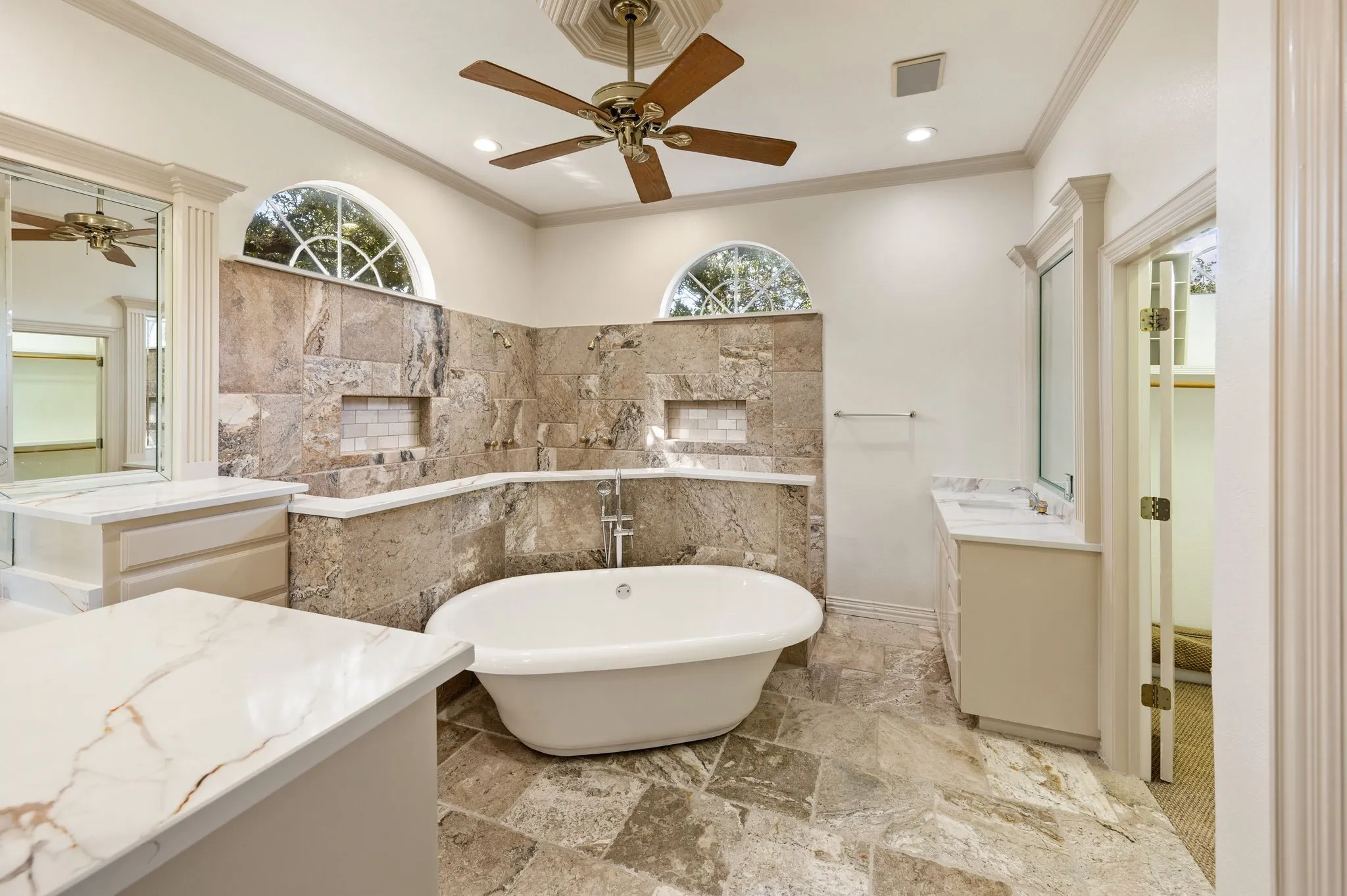 Freestanding soaking tub, stone tile flooring, dual vanities with integrated sinks, and arched transom windows