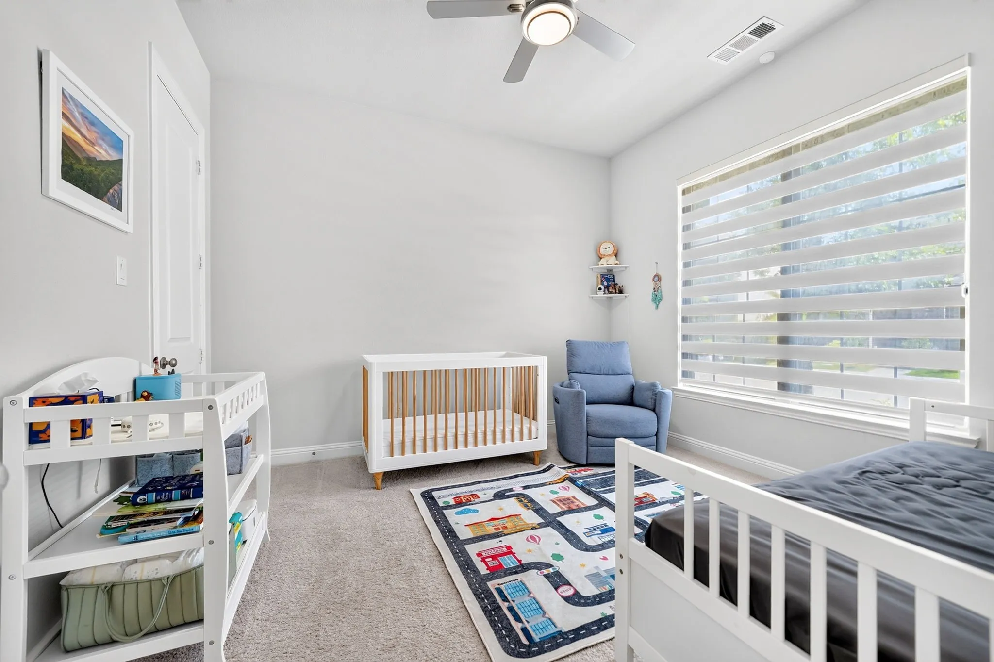 Neutral wall color, carpeted flooring, large window with horizontal blinds, overhead ceiling fan with lighting, and a white door with white trim
