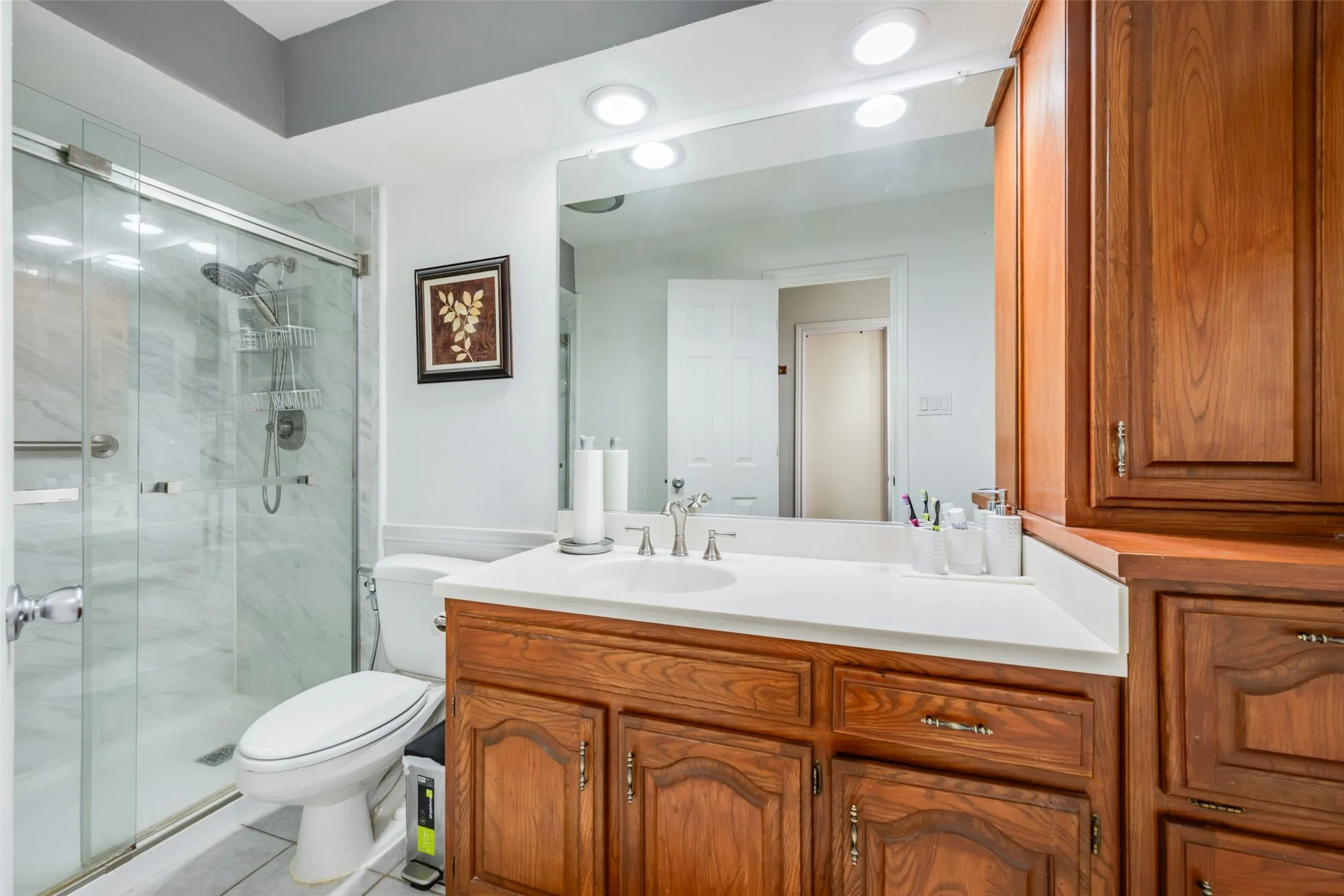Glass-enclosed shower with a marble-finish surround, wood-finish cabinetry, white countertop, and recessed lighting