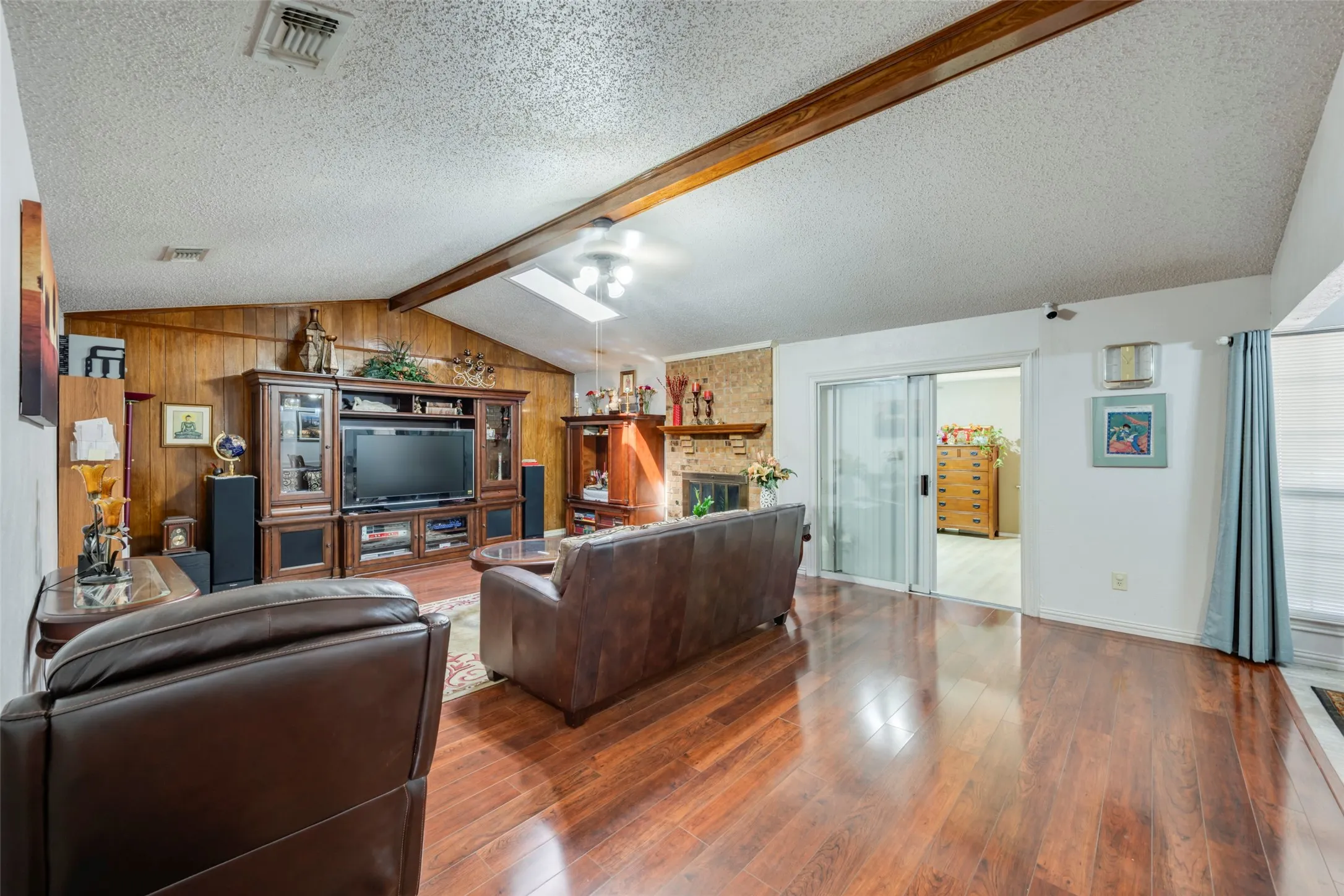 Vaulted ceiling with exposed wood beams, wood-finish flooring, wood paneling, sliding glass door, and a fireplace with a stone surround