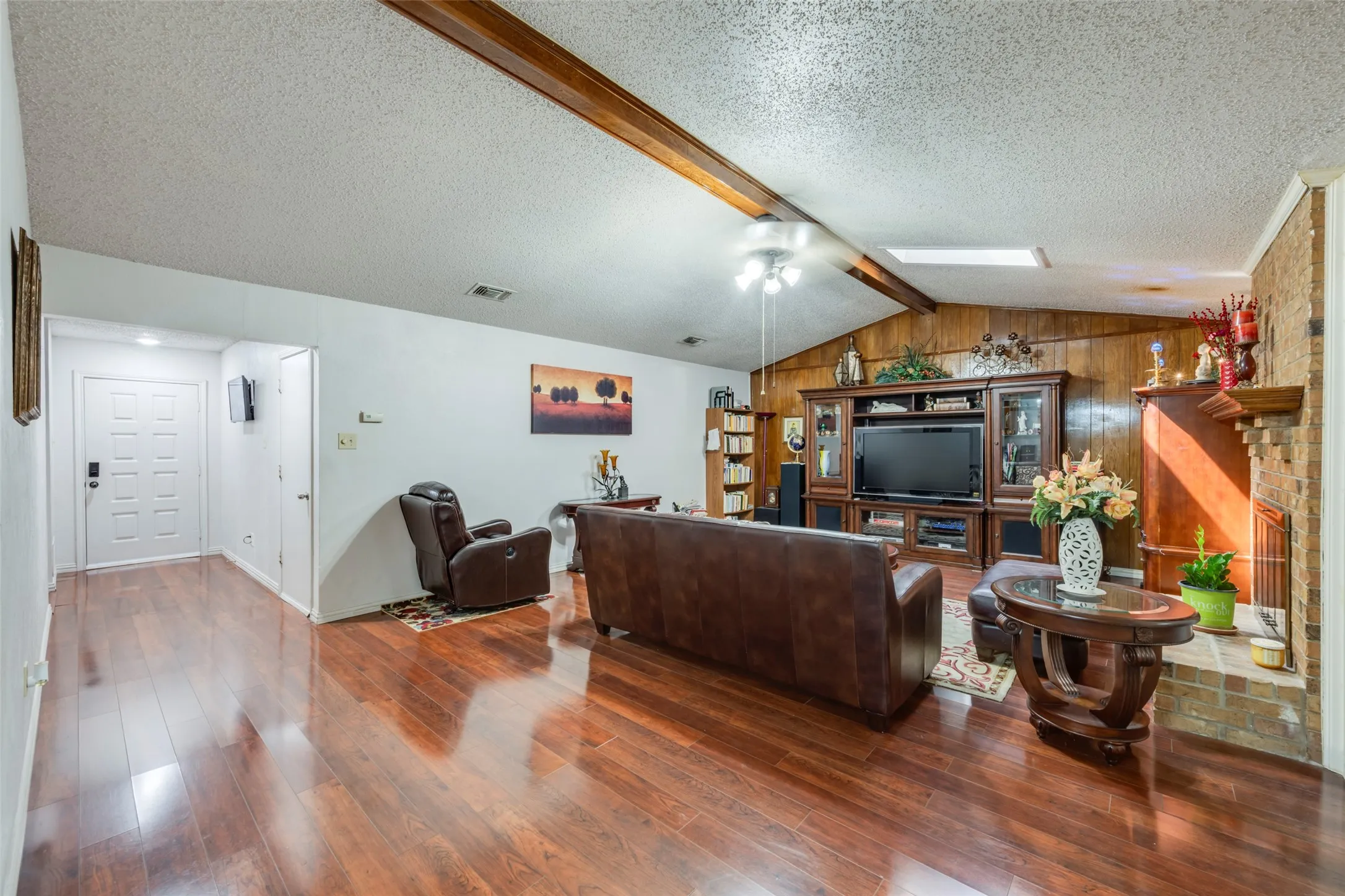 Vaulted ceiling with wood beams, wood-finish flooring, brick fireplace, and wood paneling