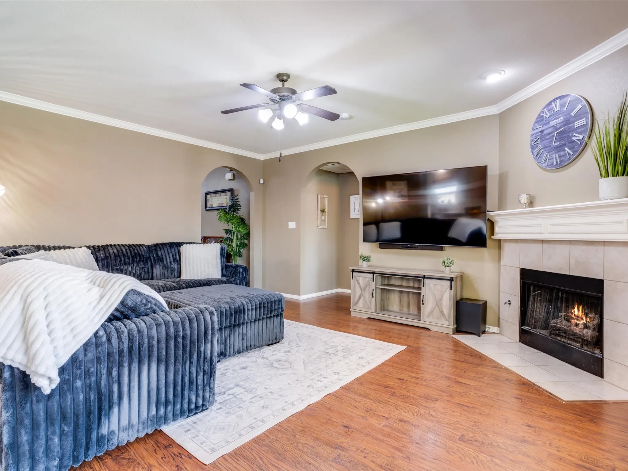 Wood-finish flooring throughout, crown molding, arched doorways, ceiling fan with integrated lighting, and a fireplace with a stone surround and mantel