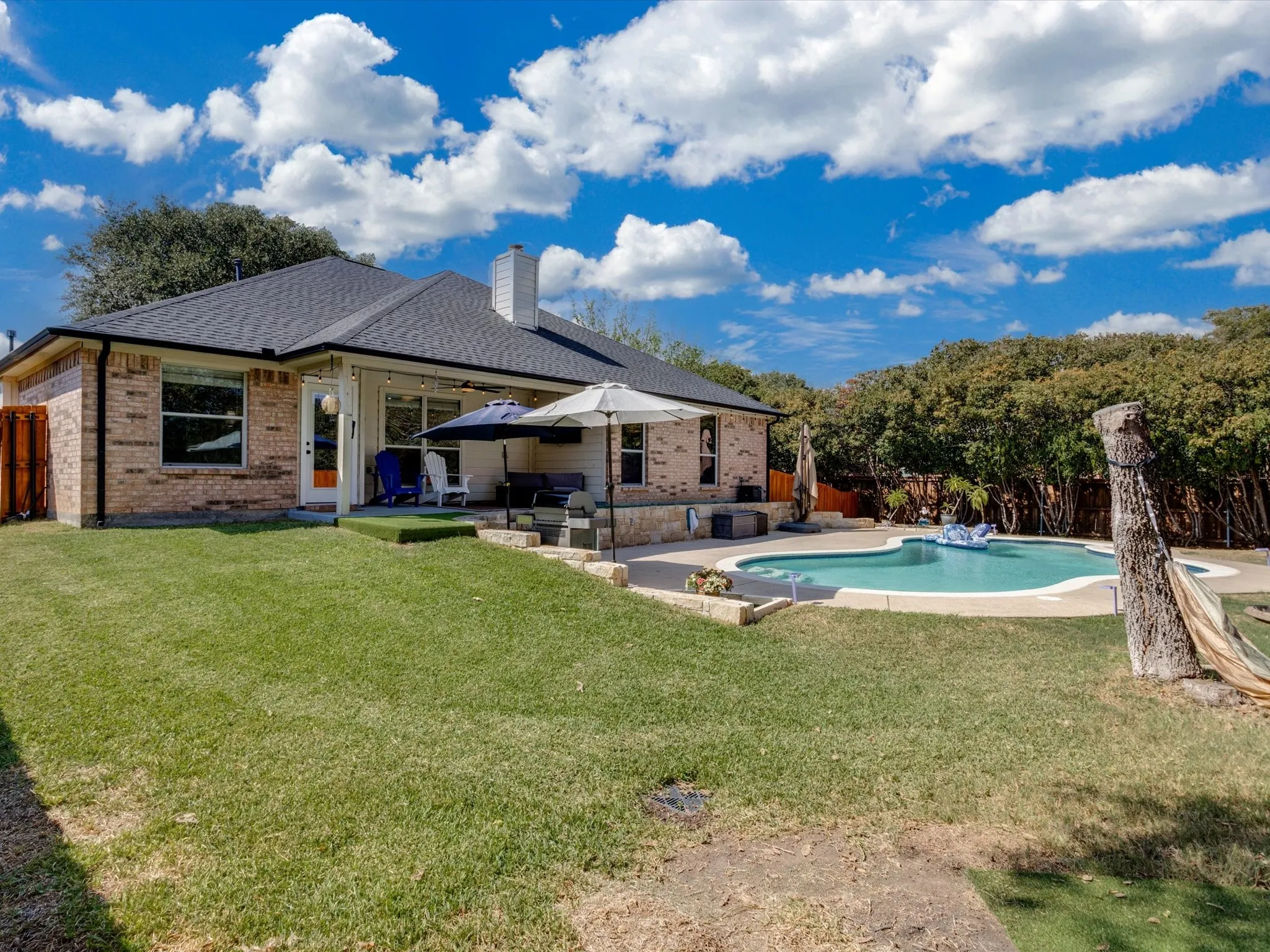 Brick exterior with a dark shingle roof, featuring a covered patio, an in-ground swimming pool, and a fenced yard with mature landscaping