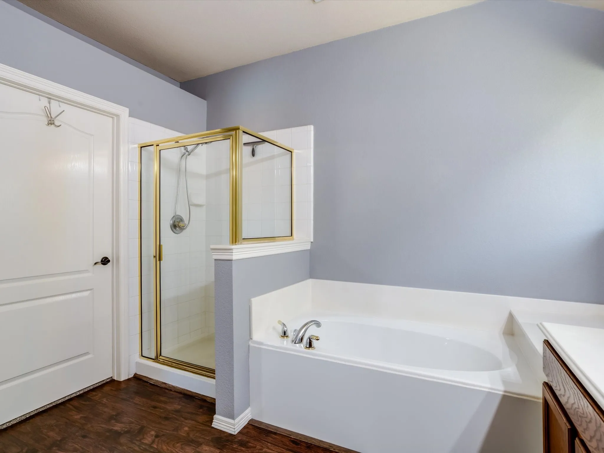 Enclosed shower with gold-tone frame, built-in bathtub, white wall tiling, wood-finish flooring, and a vanity with a light-toned countertop